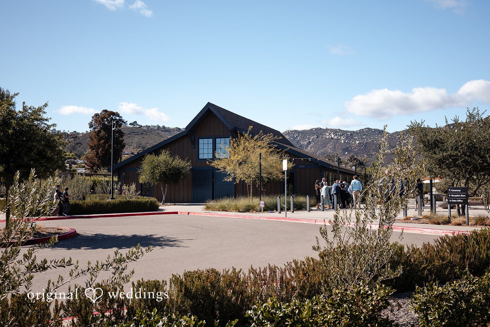 Noah + Jade Modern dark wood building with a pointed roof surrounded by green bushes and a paved walkway.