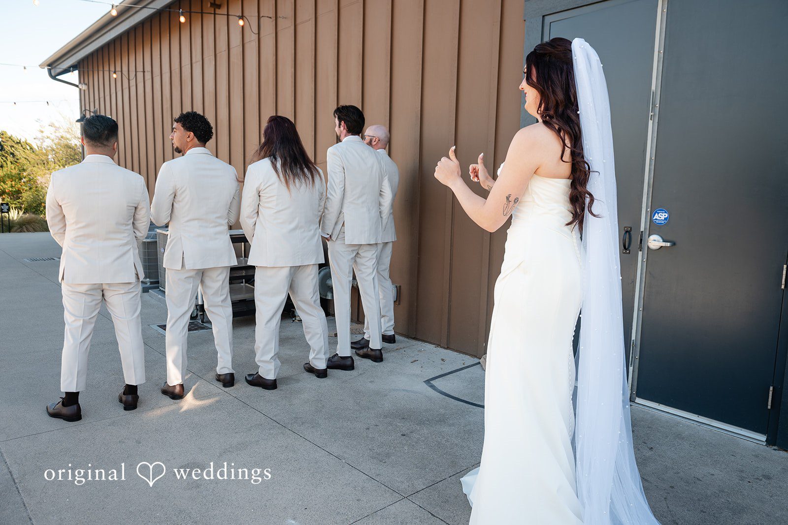 Noah + Jade A bride in a long veil standing in front of five groomsmen lined up with their backs turned.