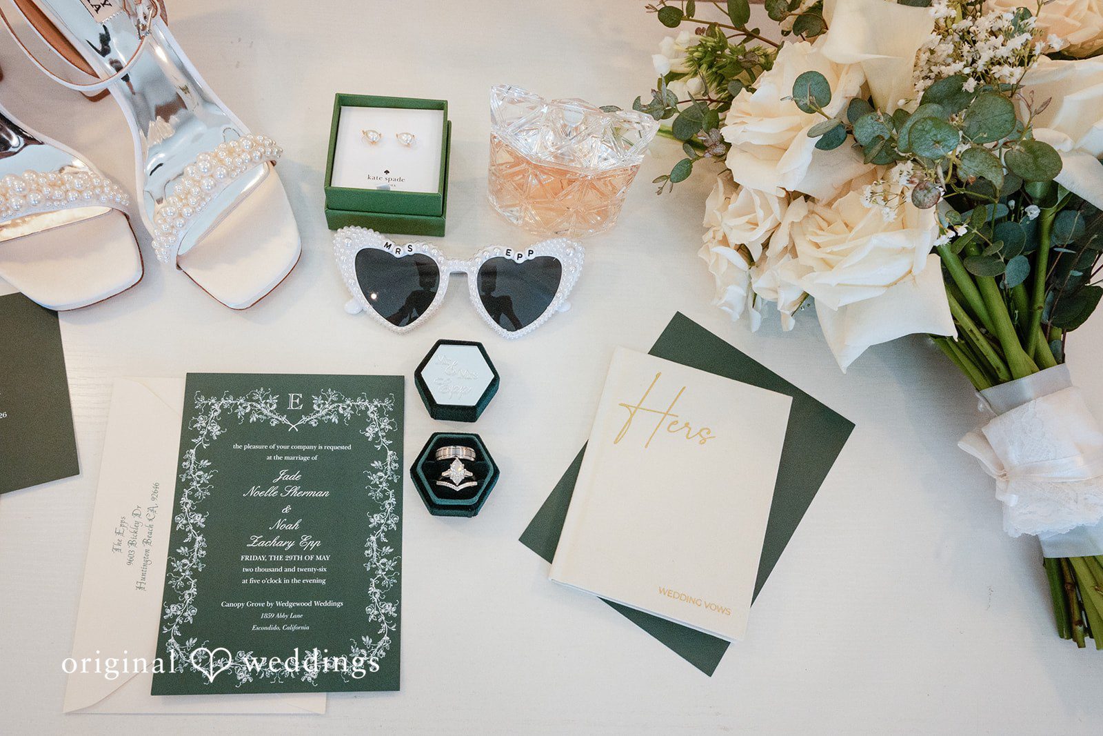 Noah + Jade Flat lay arrangement of wedding stationery, including a dark green invitation card, bridal shoes, rings, heart-shaped sunglasses, and a bouquet.
