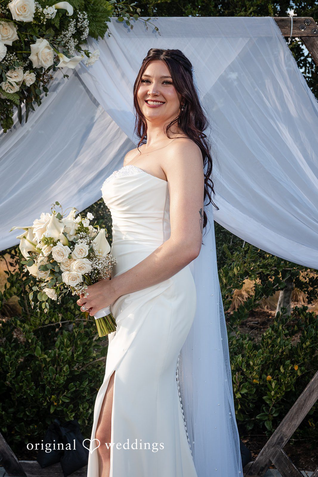 Noah + Jade Smiling bride in a fitted white wedding dress holding a bouquet in front of white draped fabric.