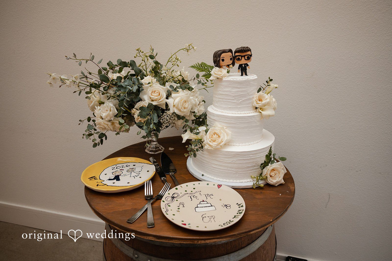 Noah + Jade Three-tiered white wedding cake decorated with flowers sitting on a round wooden table alongside small plates.