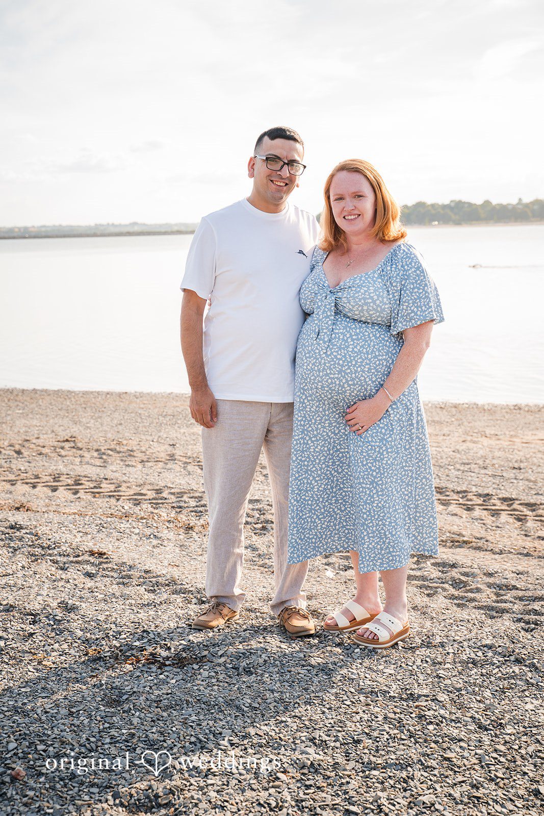 Kelly + Geoffrey Portrait of a couple standing side-by-side on a textured stone ground next to a beach line.