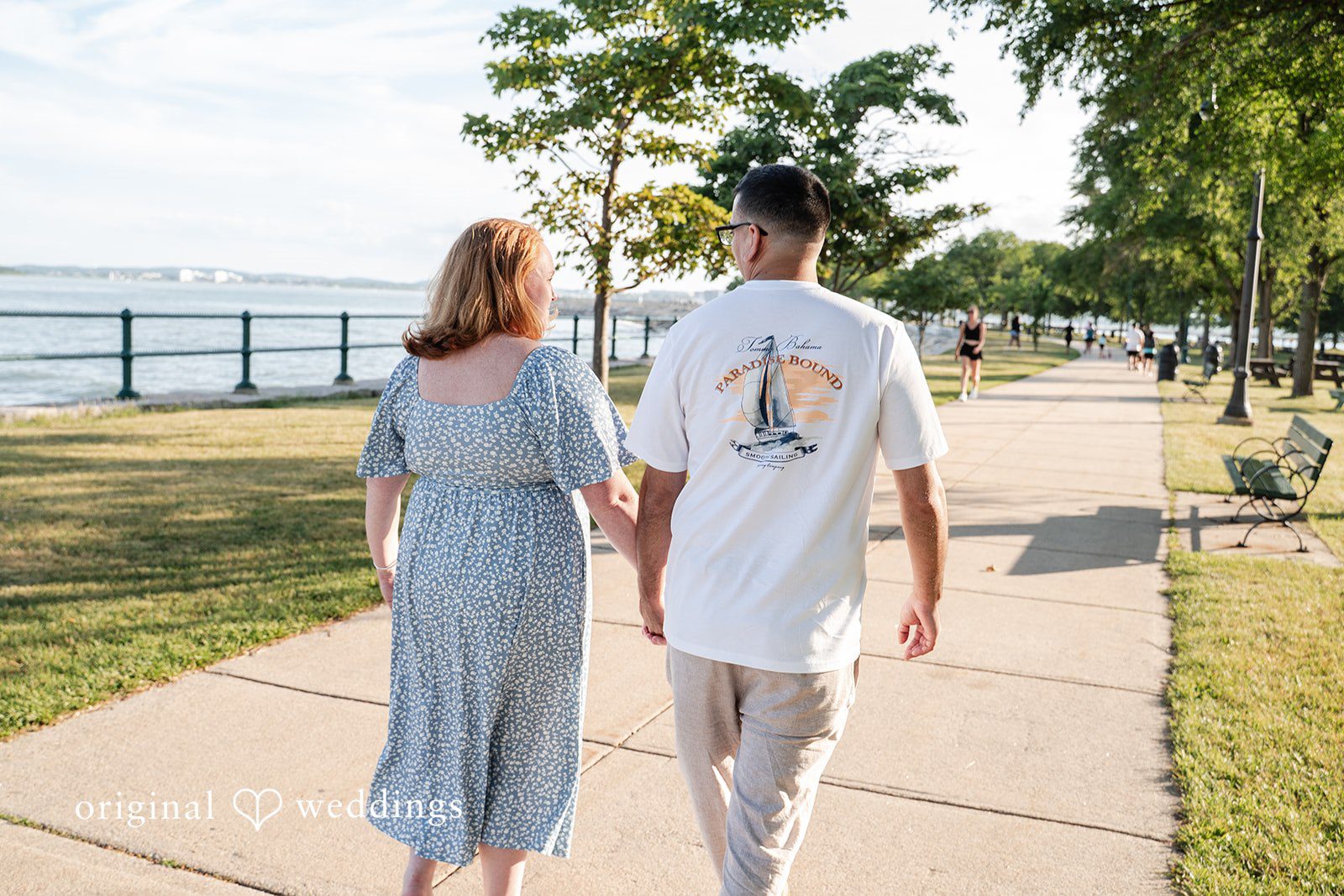 Kelly + Geoffrey A view from behind of a couple holding hands as they walk away down a sunny park path surrounded by lush trees.