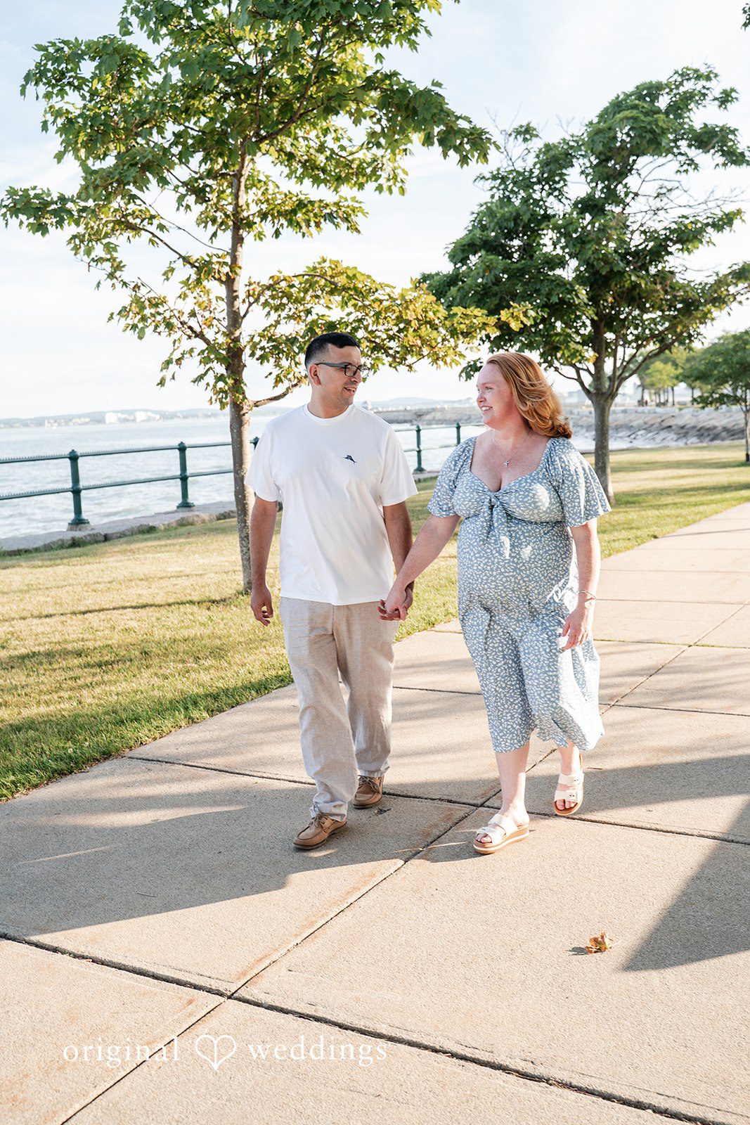 Kelly + Geoffrey A wide shot of a couple walking forward hand-in-hand along a paved concrete walkway lined with green trees.