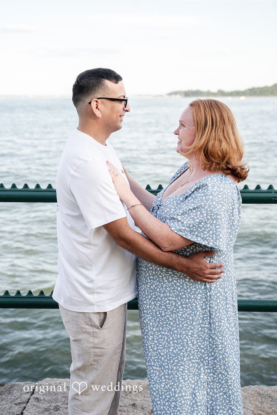 Kelly + Geoffrey A medium shot of a couple standing face-to-face, smiling and looking lovingly into each other's eyes by a waterfront railing.