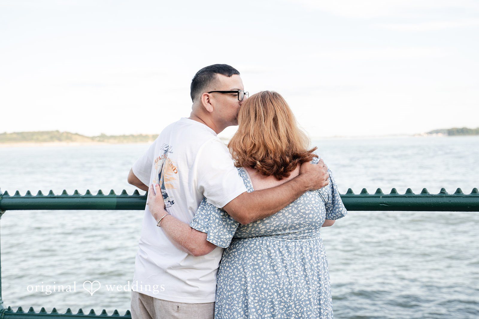 Kelly + Geoffrey A couple embracing tightly by a waterfront fence, with the man kissing the top of the woman's head from behind.