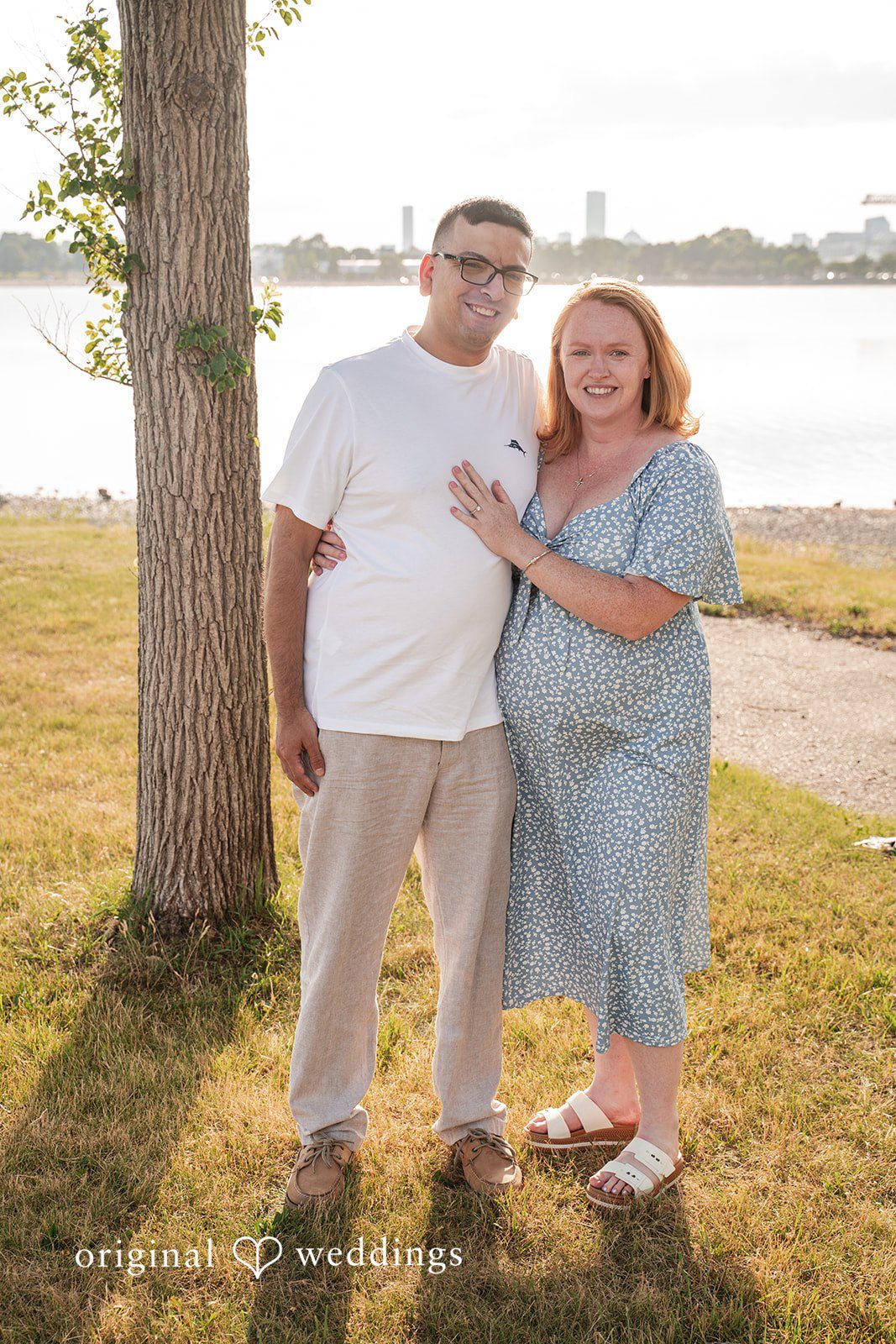 Kelly + Geoffrey Portrait of a couple standing together on a grassy lawn next to a tree trunk, smiling directly at the camera.