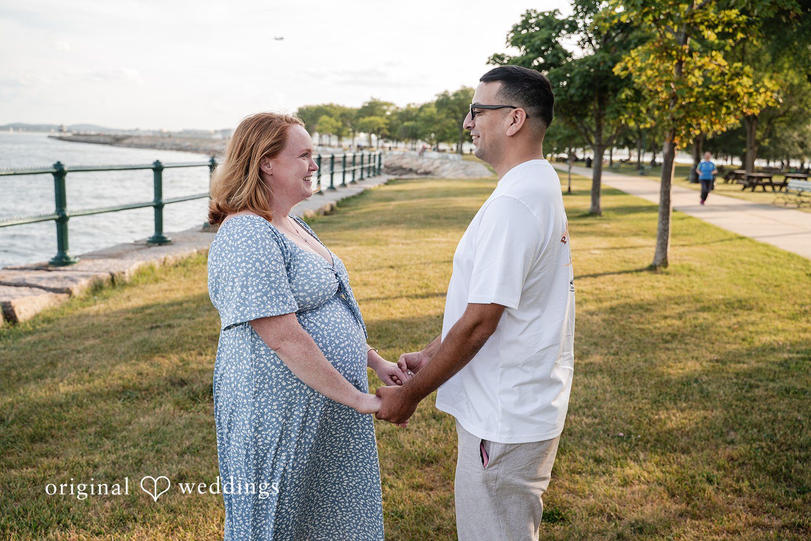 Kelly + Geoffrey A couple facing each other and holding hands while standing on a grassy path lined with trees and a fence.