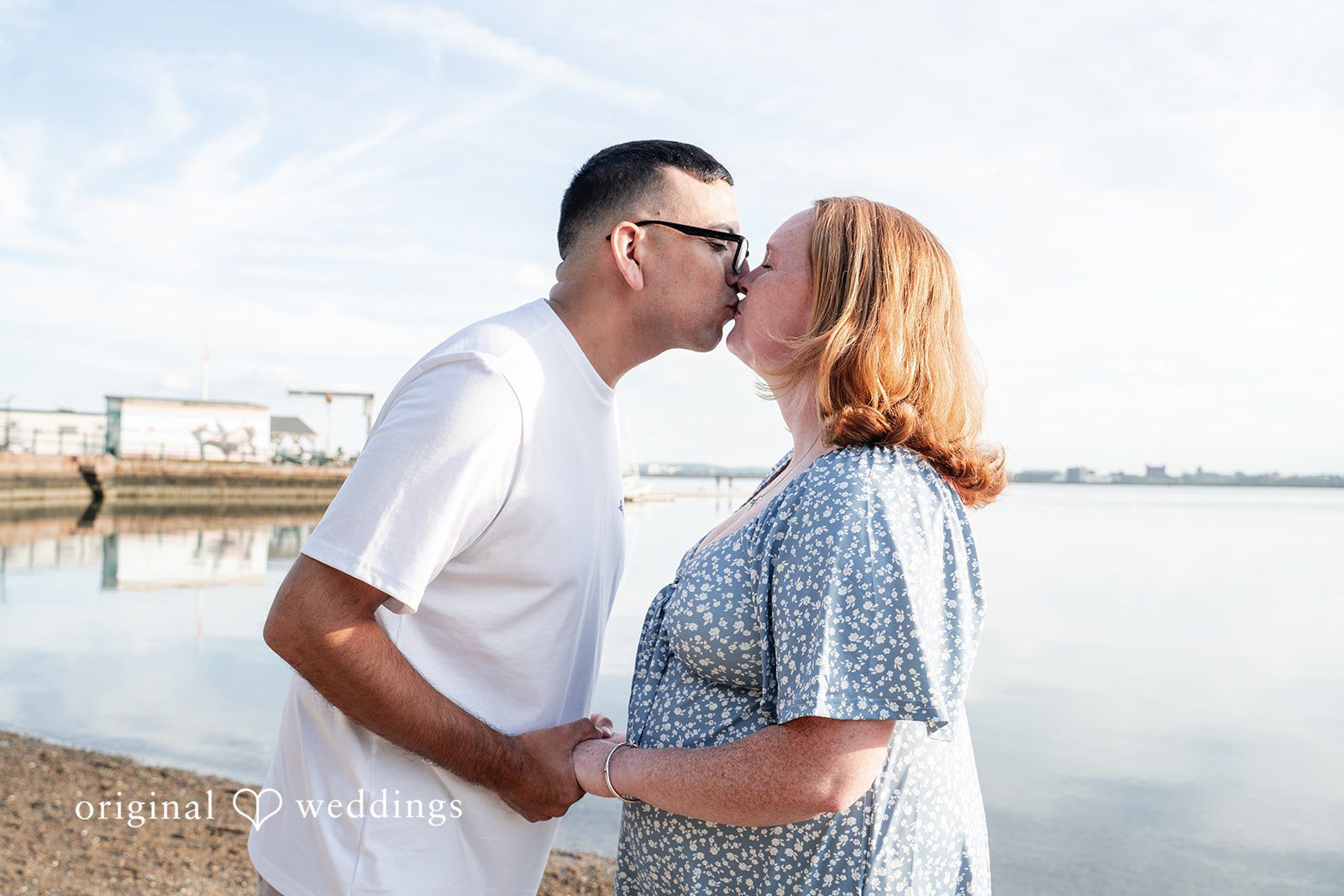 Kelly + Geoffrey A romantic profile shot of a couple sharing a sweet kiss on the lips by a sunny waterfront.