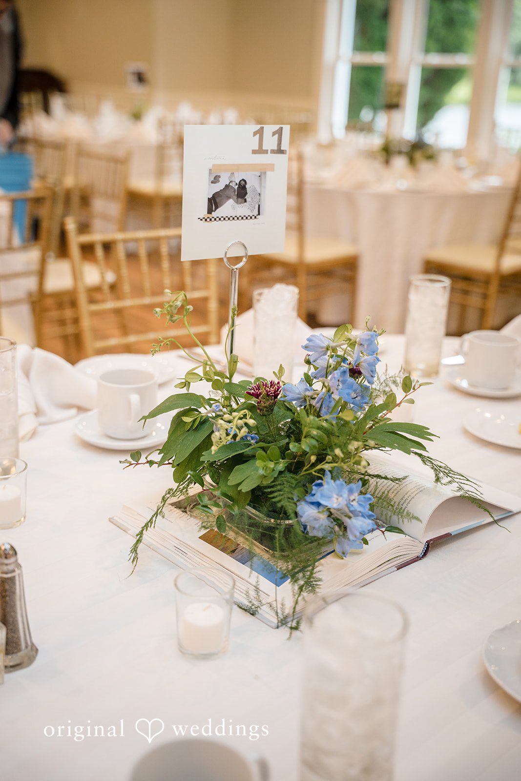 Meghan + Rowan A round wedding reception table decorated with a centerpiece of fresh blue and white flowers mixed with green foliage.