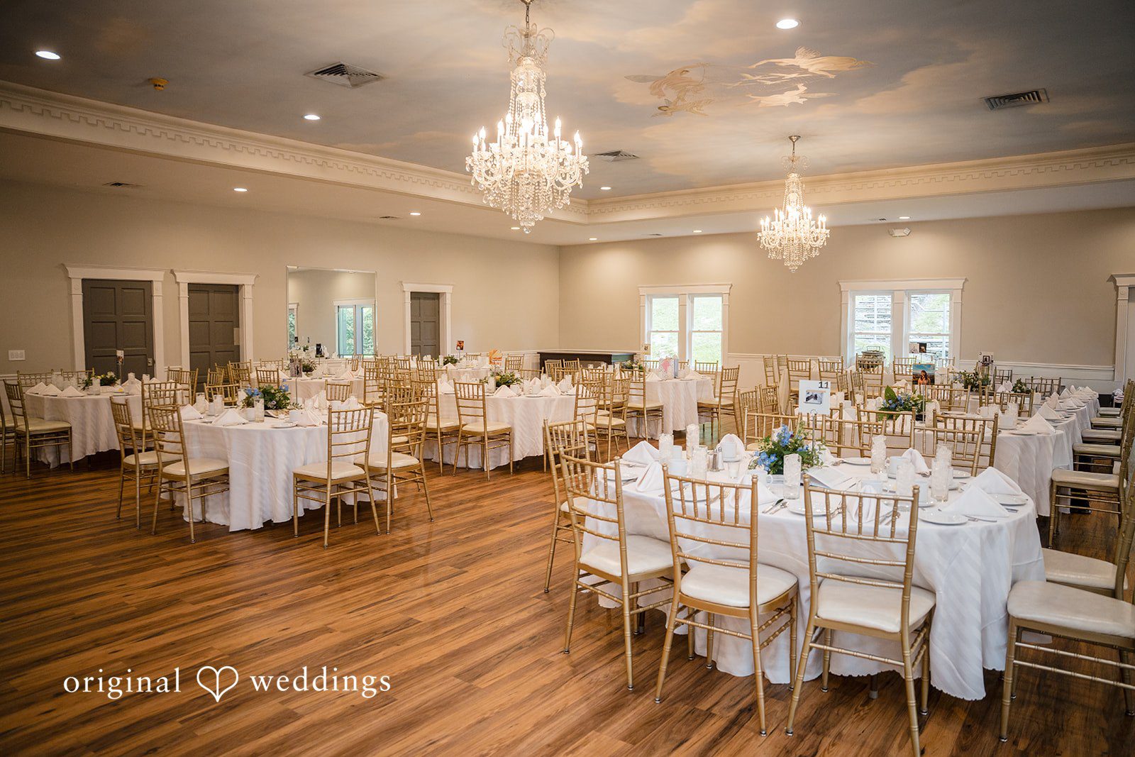 Meghan + Rowan Wedding reception venue featuring round dining tables, elegant white chairs, and crystal chandeliers hanging from the ceiling.