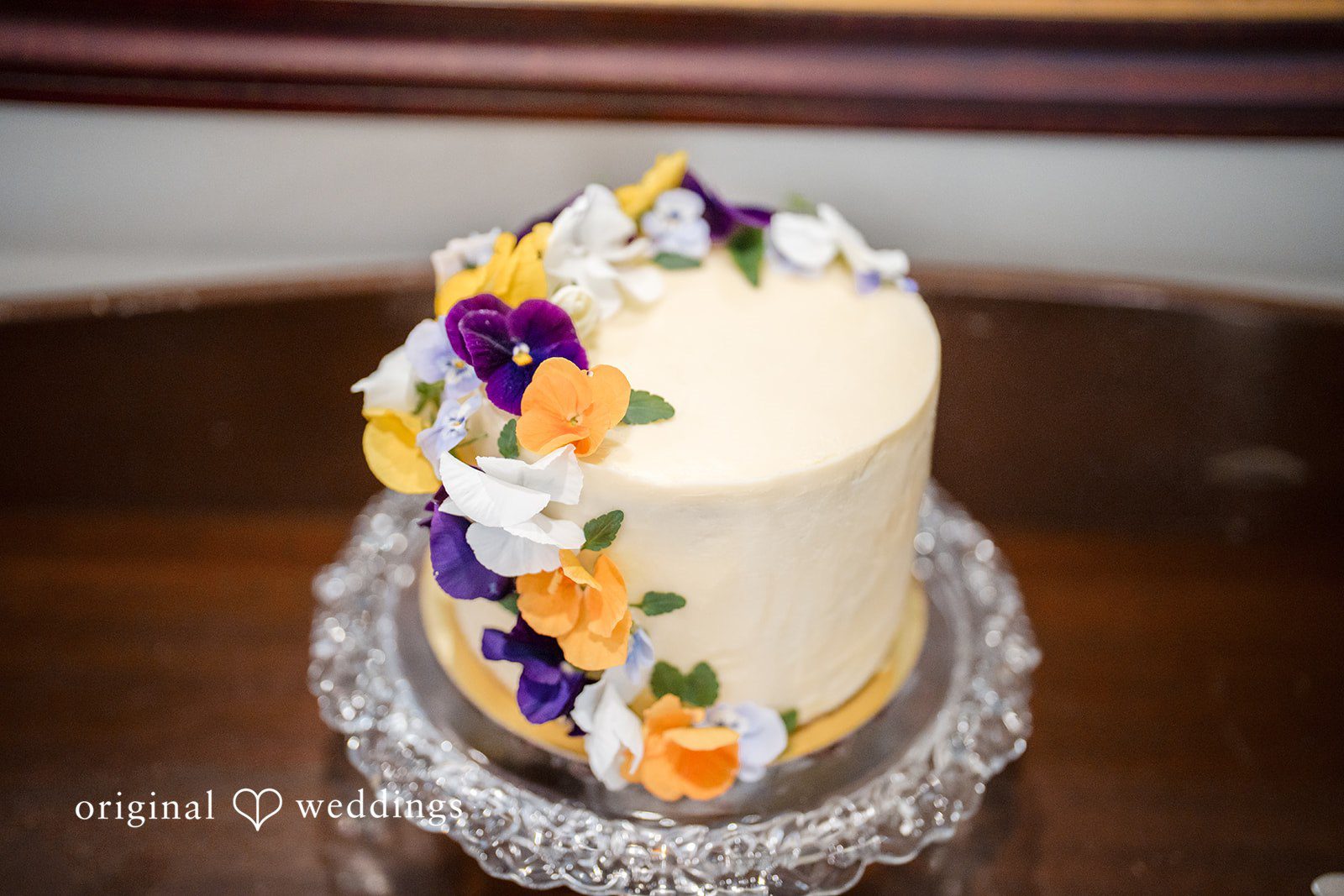 Meghan + Rowan A small white frosted wedding cake decorated with edible orange and purple flower petals.