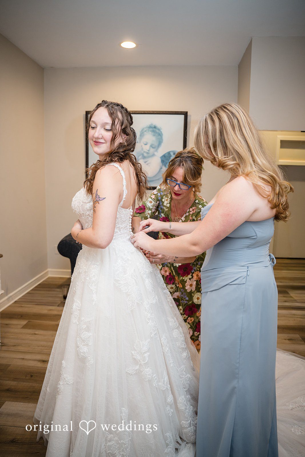 Meghan + Rowan A woman in a grey dress helping a bride adjust the back of her white wedding gown.