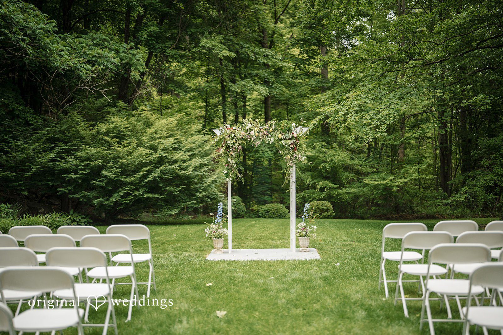 Meghan + Rowan An empty wedding ceremony setup with white chairs on grass facing a simple wooden archway.