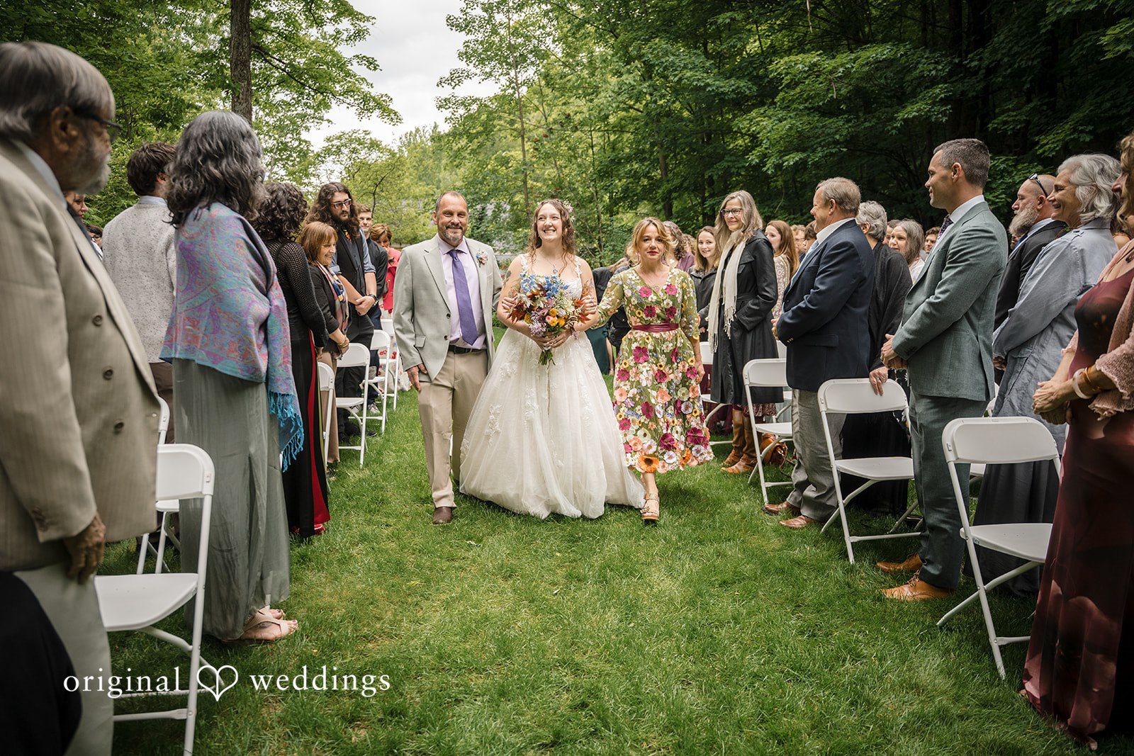 Meghan + Rowan A bride walking down a grassy ceremony aisle lined with wedding guests seated in white chairs.