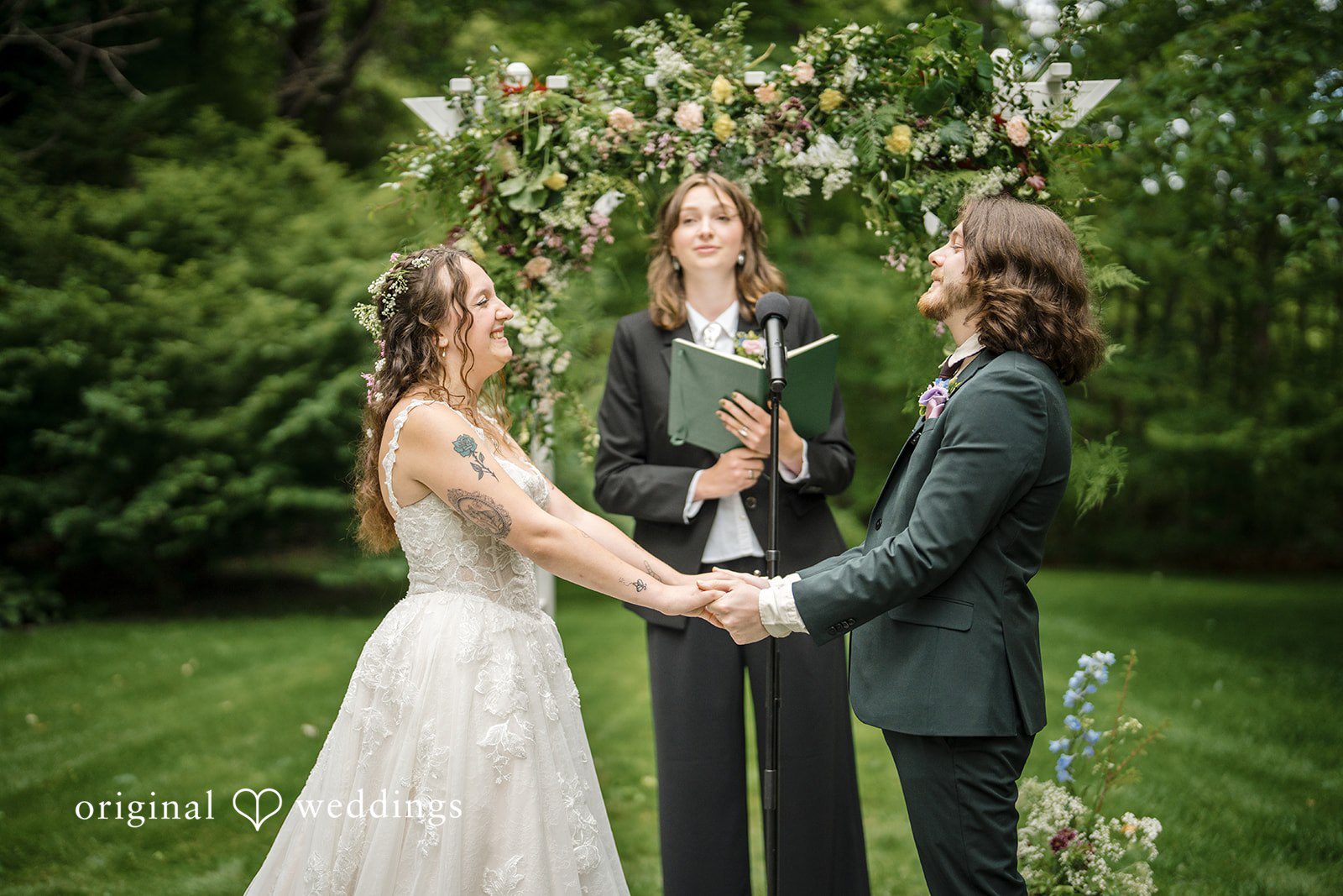 Meghan + Rowan A bride and groom holding hands under a floral archway while an officiant conducts their ceremony.
