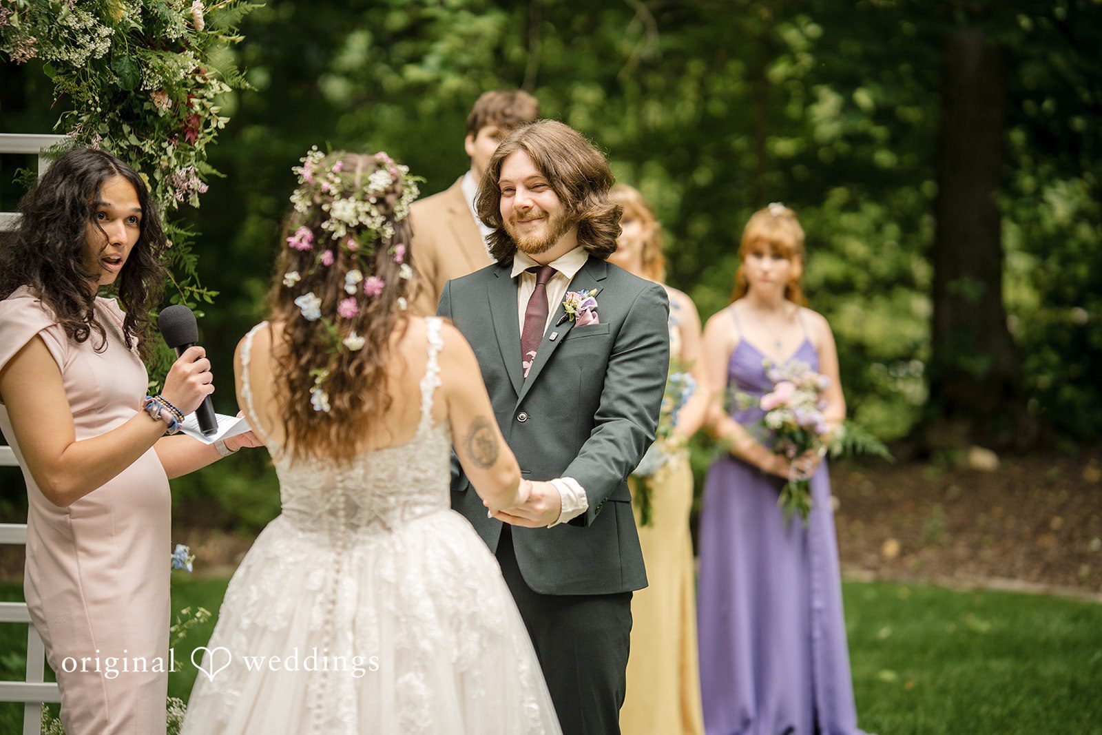 Meghan + Rowan A bride and groom holding hands at the altar, viewed from behind the bride, with their bridal party standing nearby.