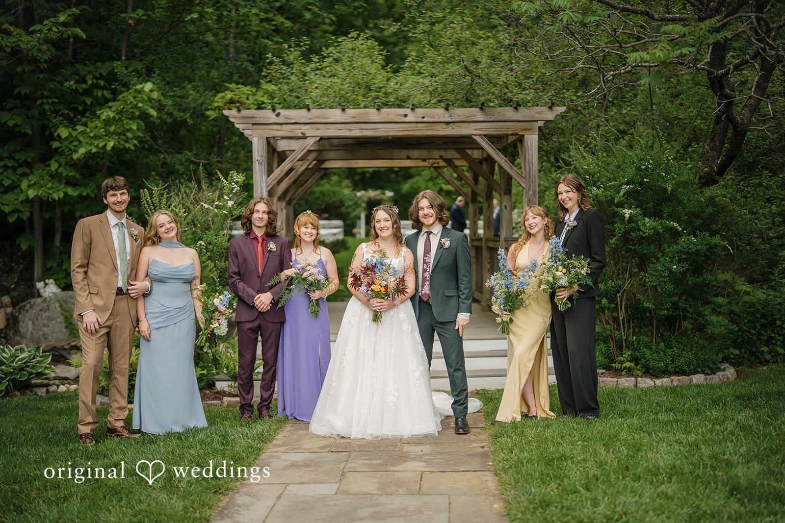 Meghan + Rowan A wedding party poses together under a rustic wooden pergola structure surrounded by green trees.