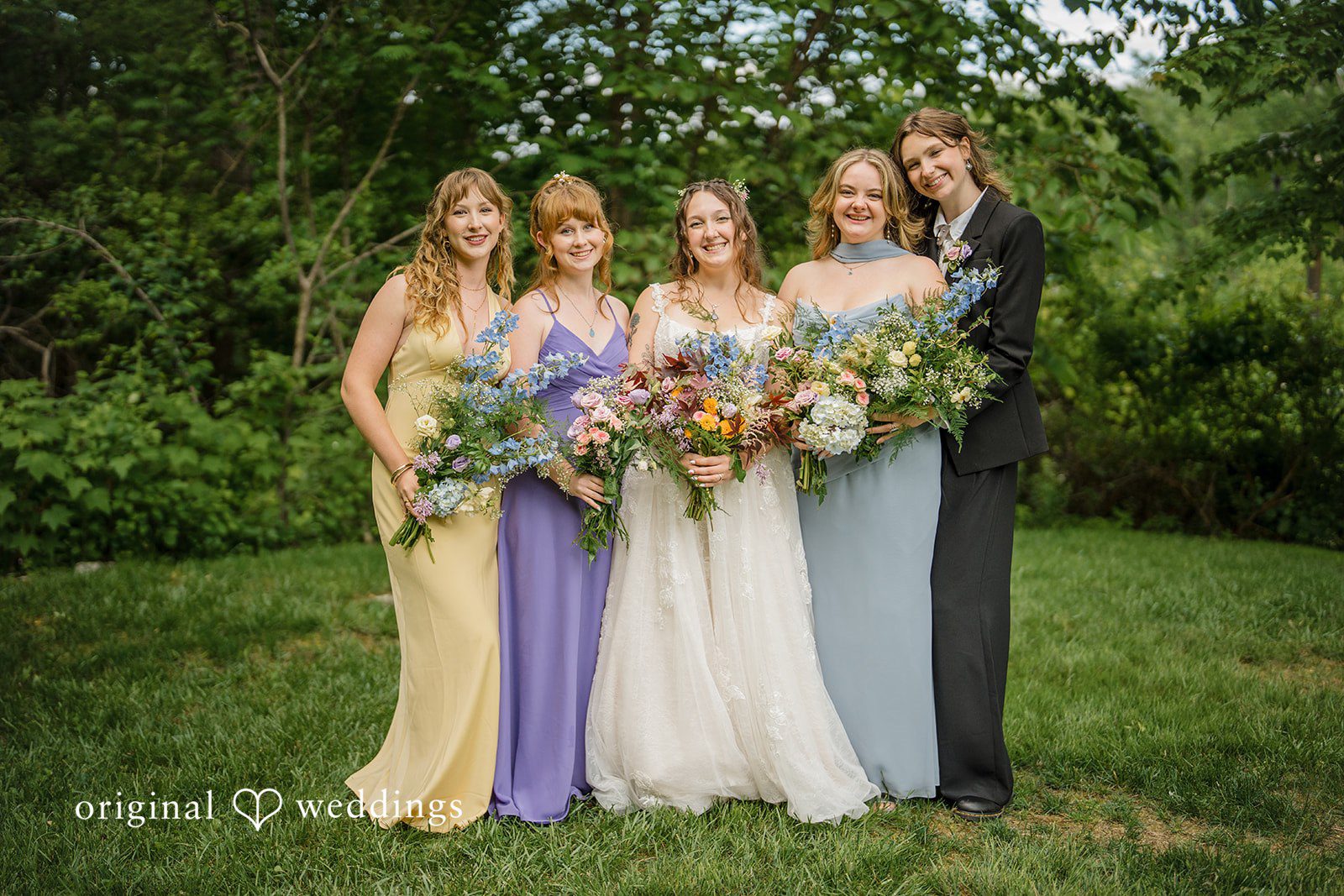 Meghan + Rowan A bride posing on a lawn with three bridesmaids wearing mismatched pastel dresses in shades of purple, yellow, and blue.