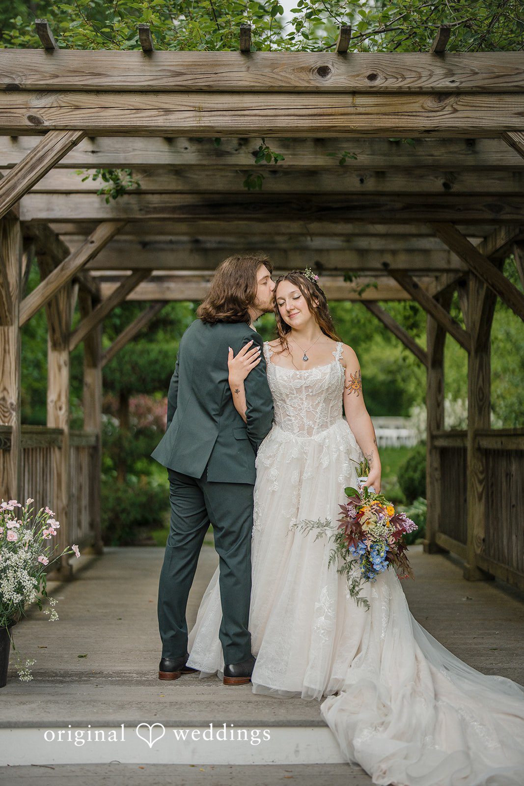 Meghan + Rowan A bride and groom posing together under a large wooden pergola structure, with the bride's gown train fully extended.