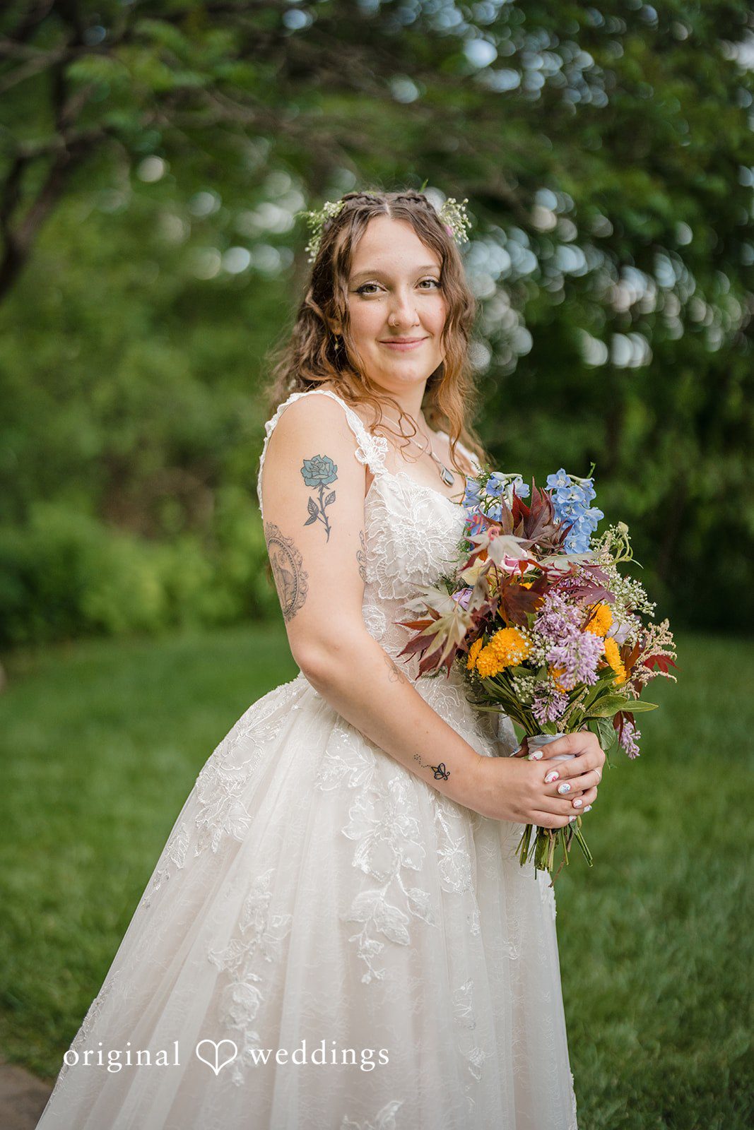 Meghan + Rowan A bride in a white gown posing with a vibrant, multi-colored floral bouquet against a lush green backdrop.