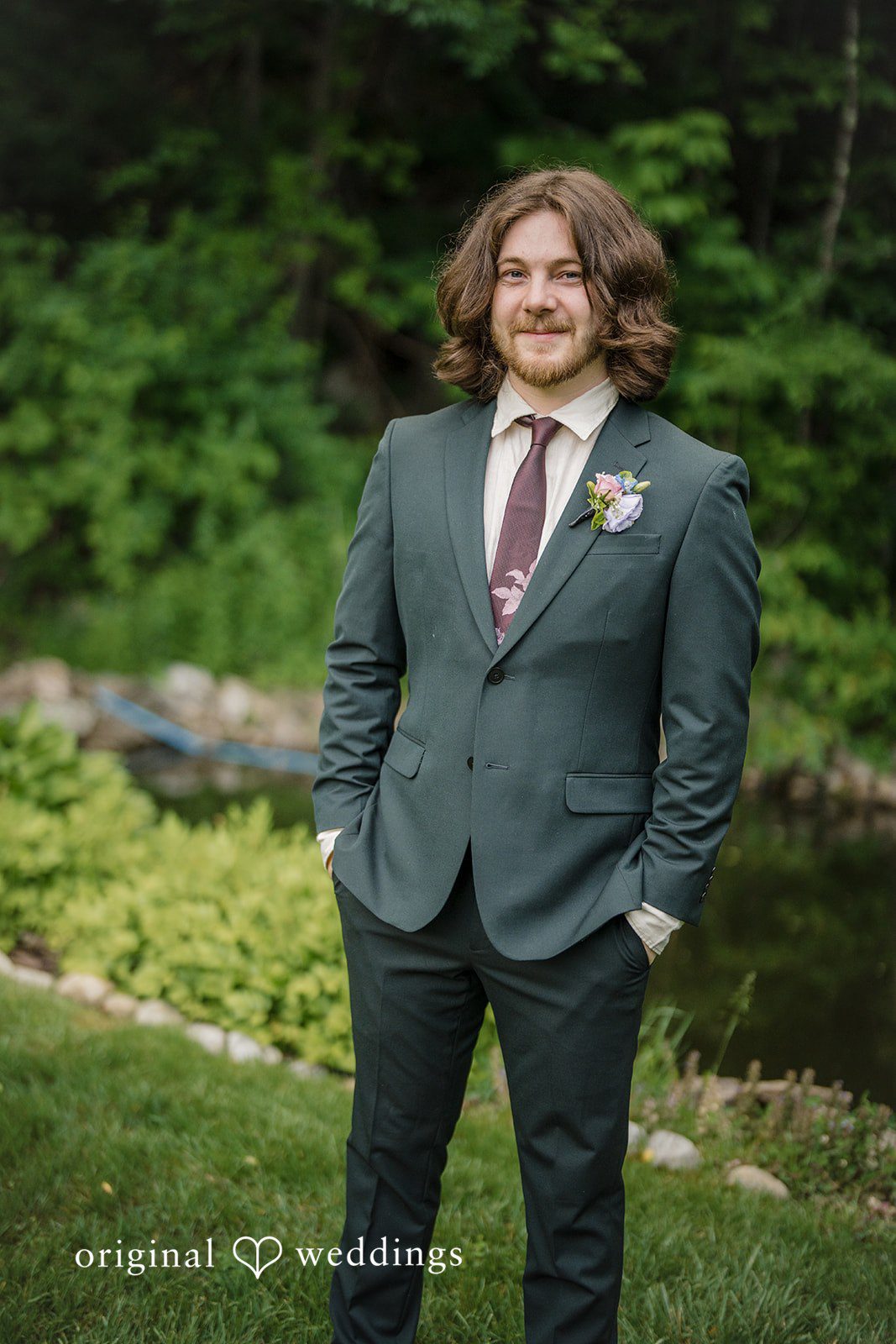 Meghan + Rowan A groom with wavy hair wearing a dark grey suit and a red tie, standing in a natural setting.