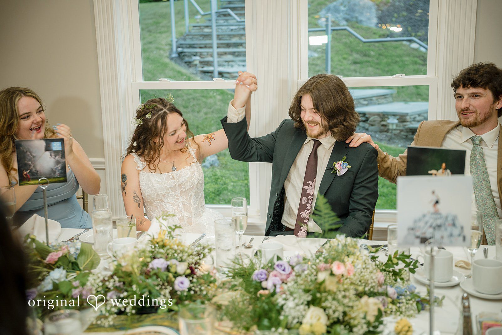 Meghan + Rowan A groom raises his fist in celebration while sitting next to his bride at a decorated reception head table.