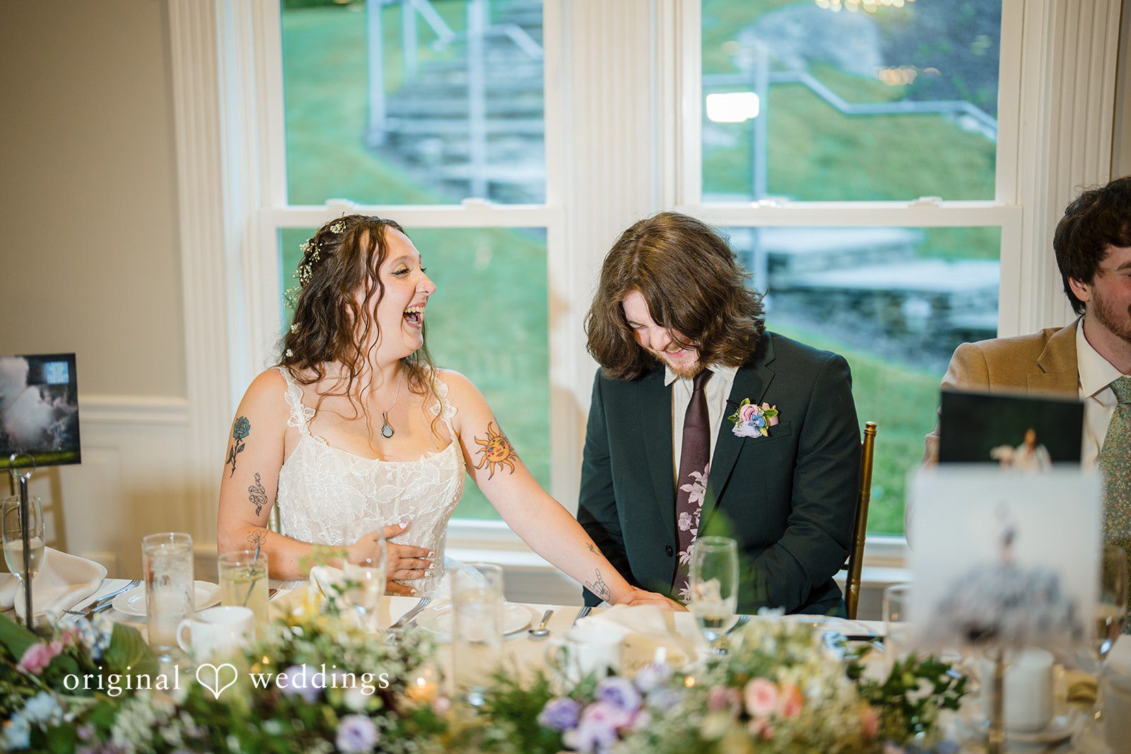 Meghan + Rowan A bride and groom smile and interact with guests while seated at a reception dining table.