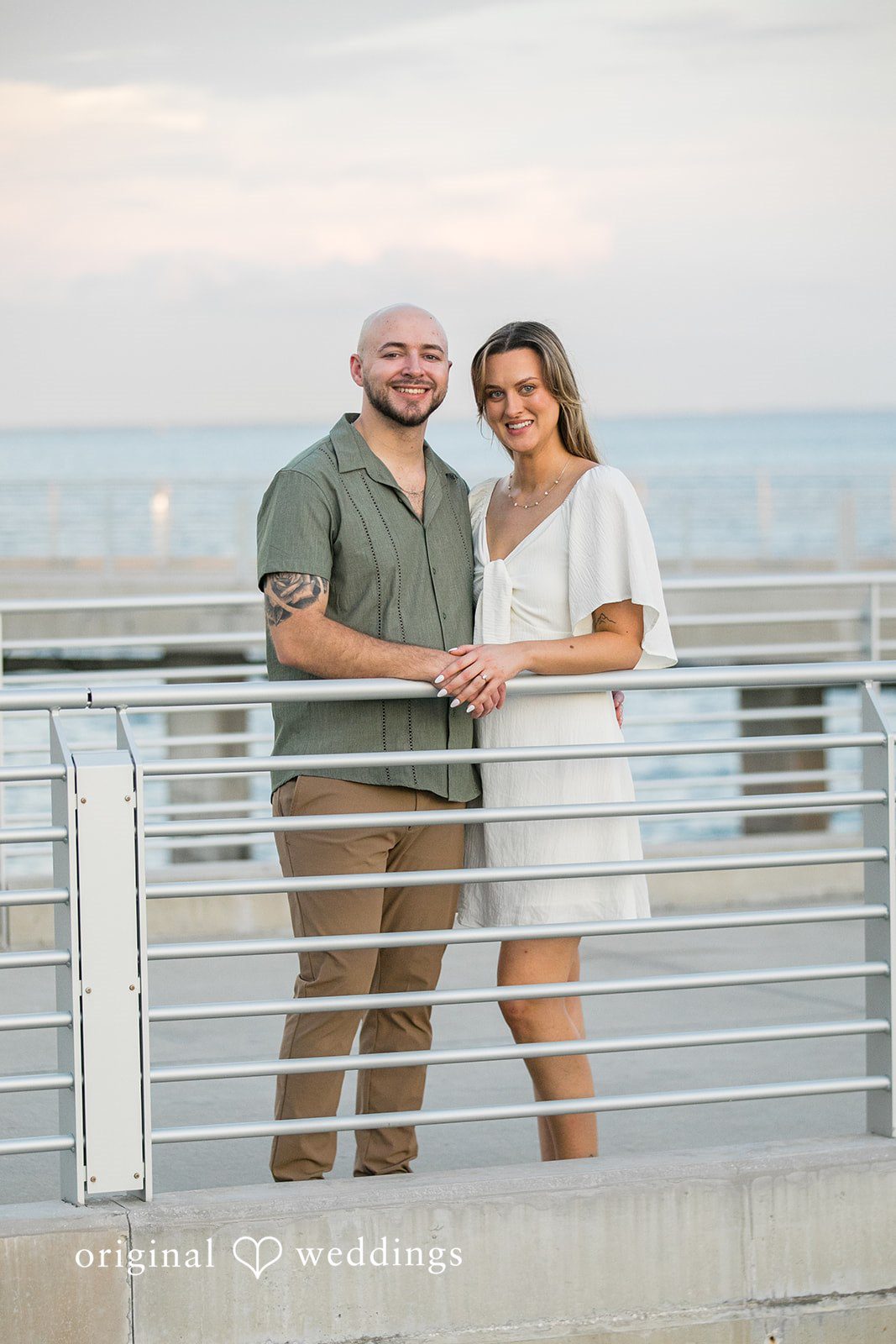 Lauren + Miles A couple standing side-by-side and smiling behind a metal railing.