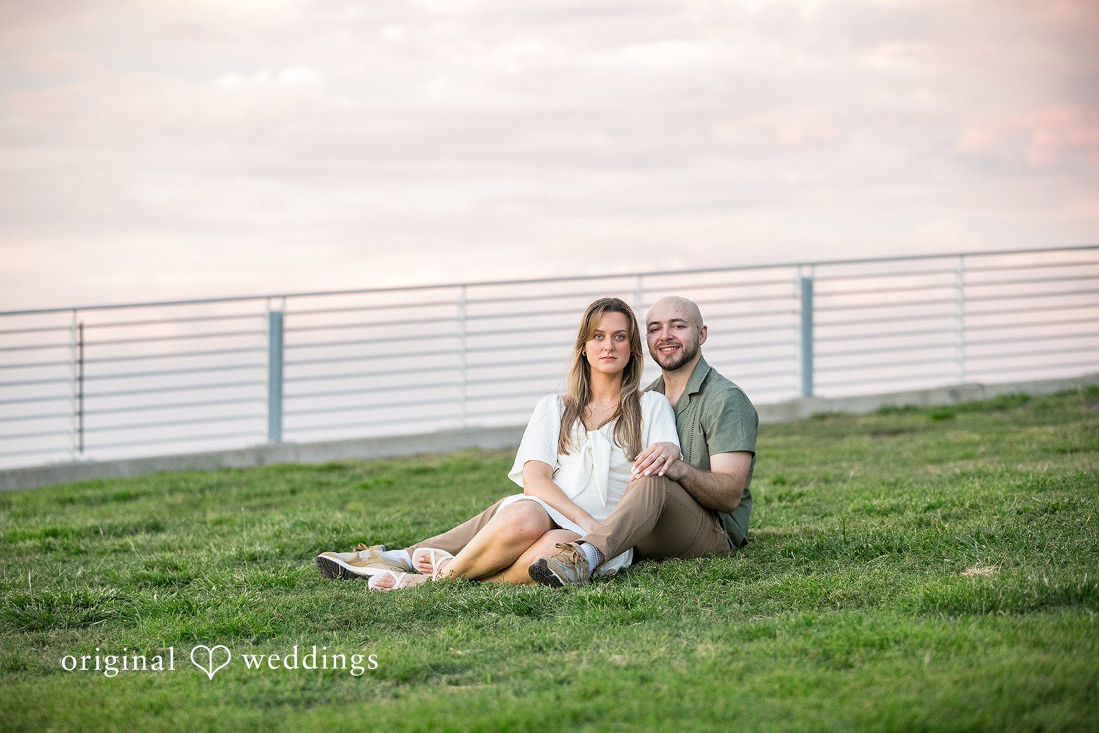 Lauren + Miles A couple sitting together on a grassy hill near a metal railing.