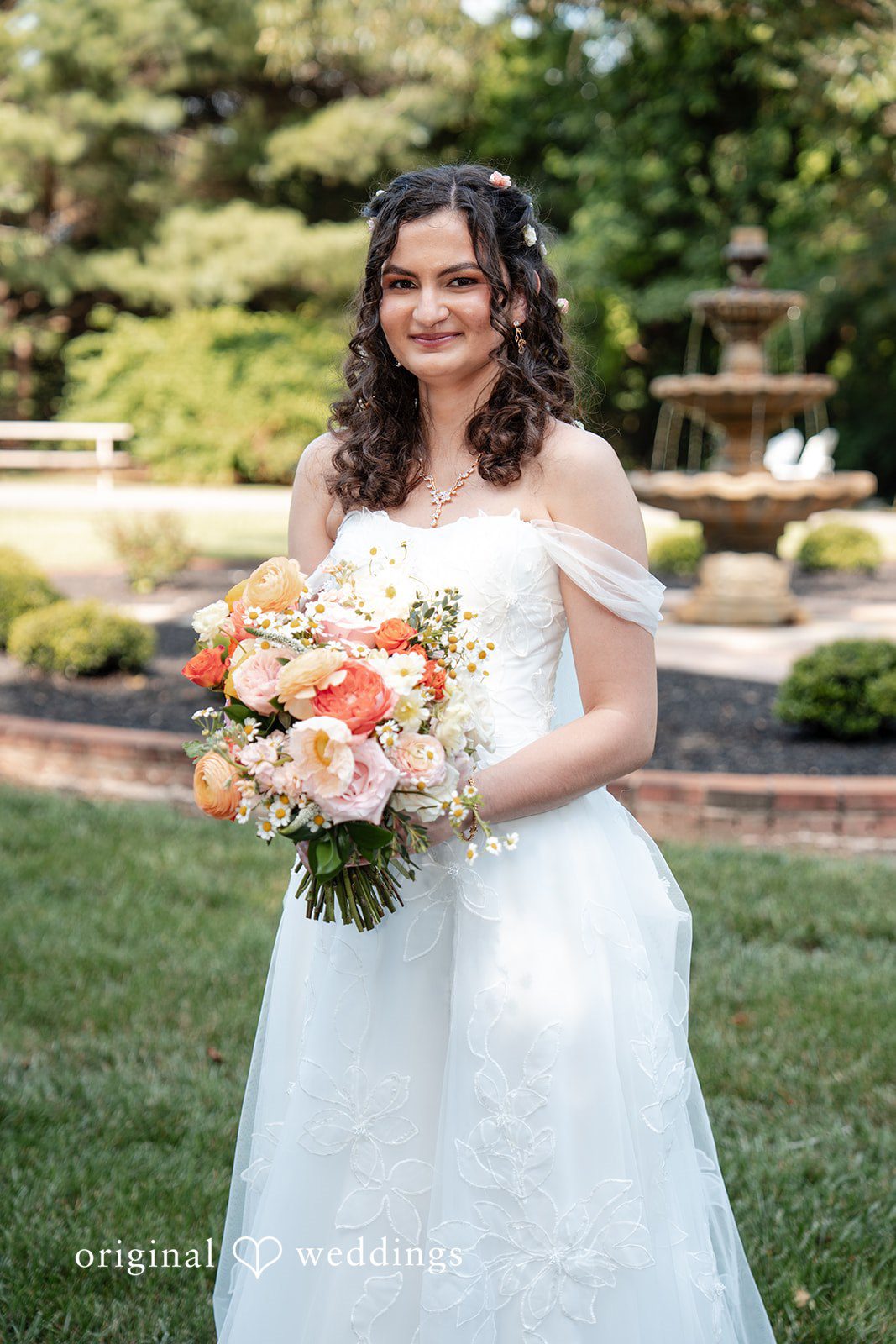 Conner + Ashley A smiling bride standing on a green lawn holding a bouquet of peach and white flowers.