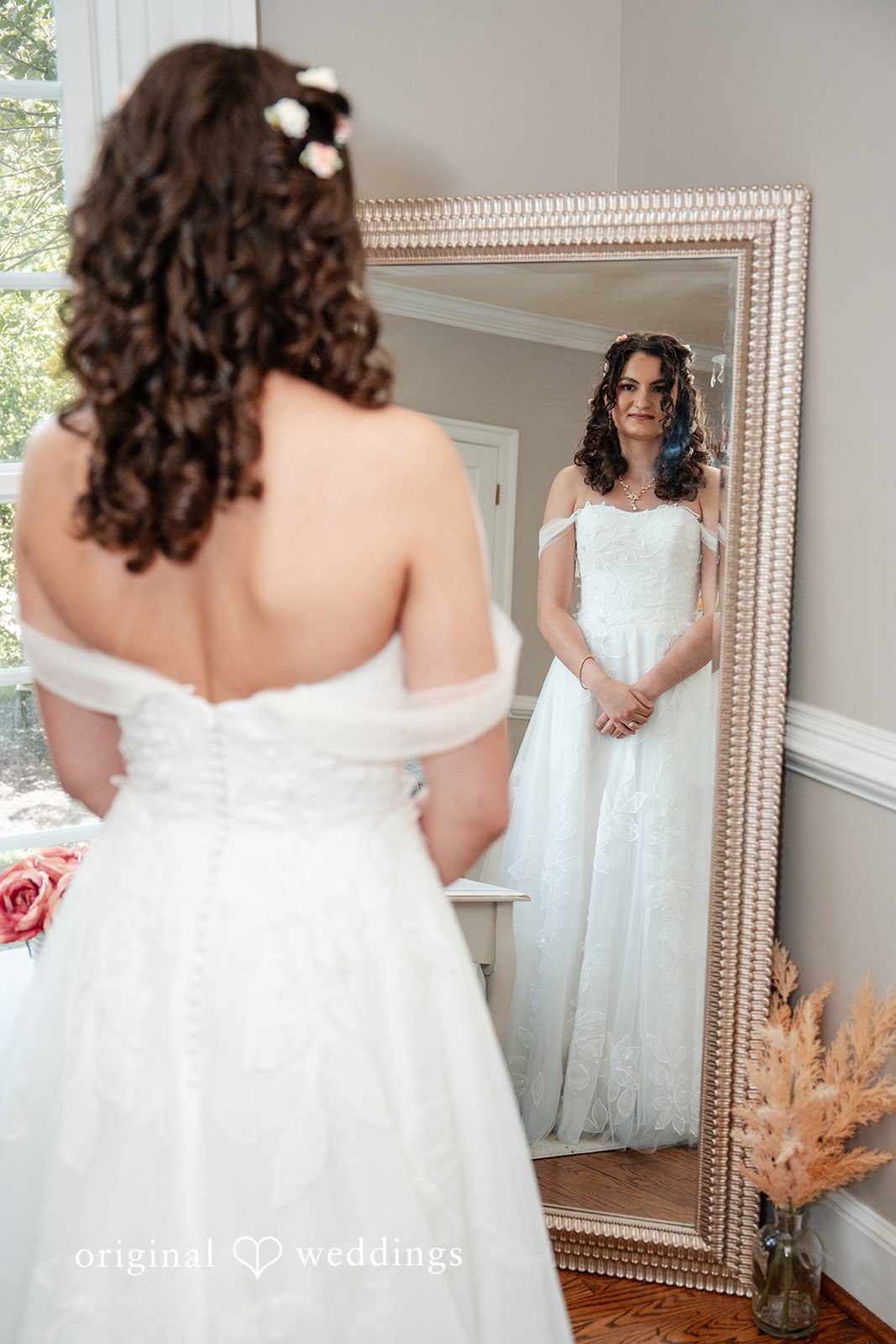 Conner + Ashley A bride looking into a large ornate mirror while wearing an off-the-shoulder wedding gown.