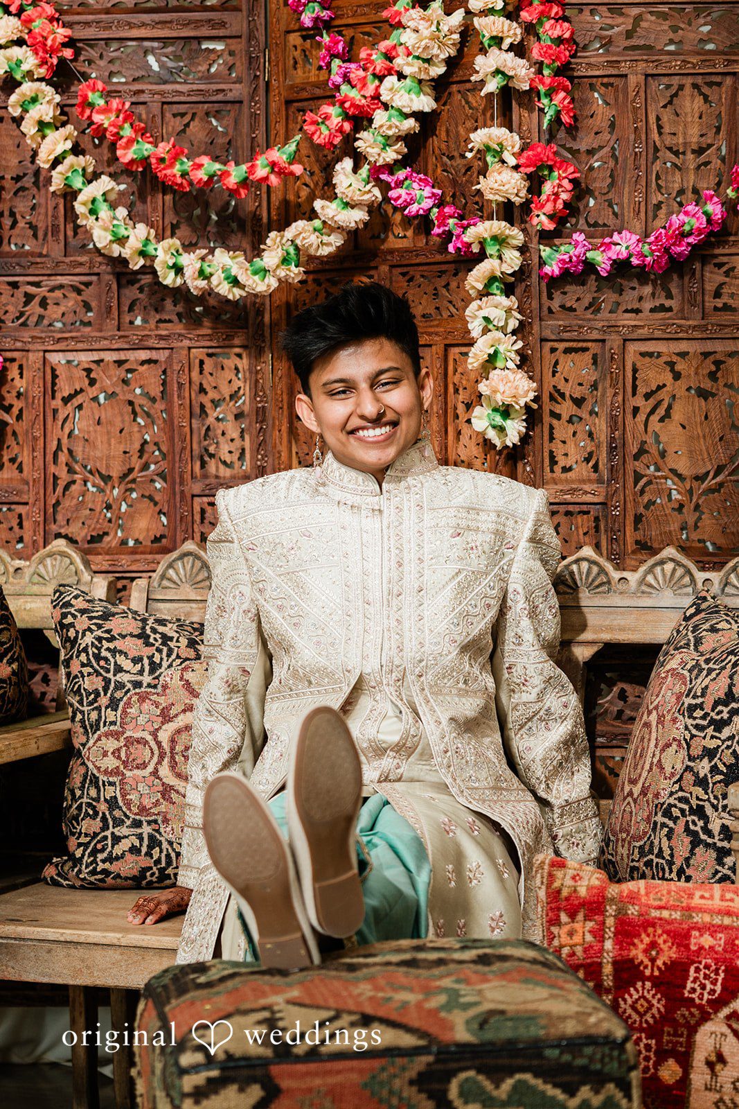 Sonya + Akshaya A groom sitting on a decorated platform with floral garlands hanging behind him.