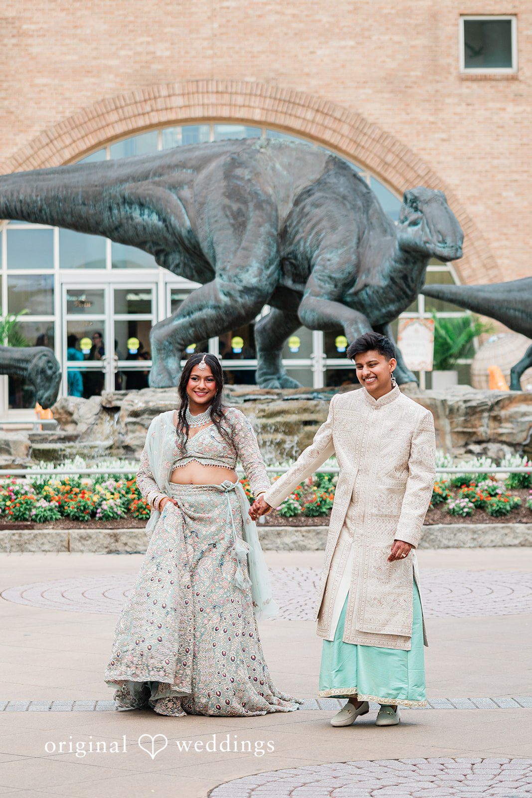 Sonya + Akshaya A bride and groom holding hands while walking in front of a dinosaur statue.