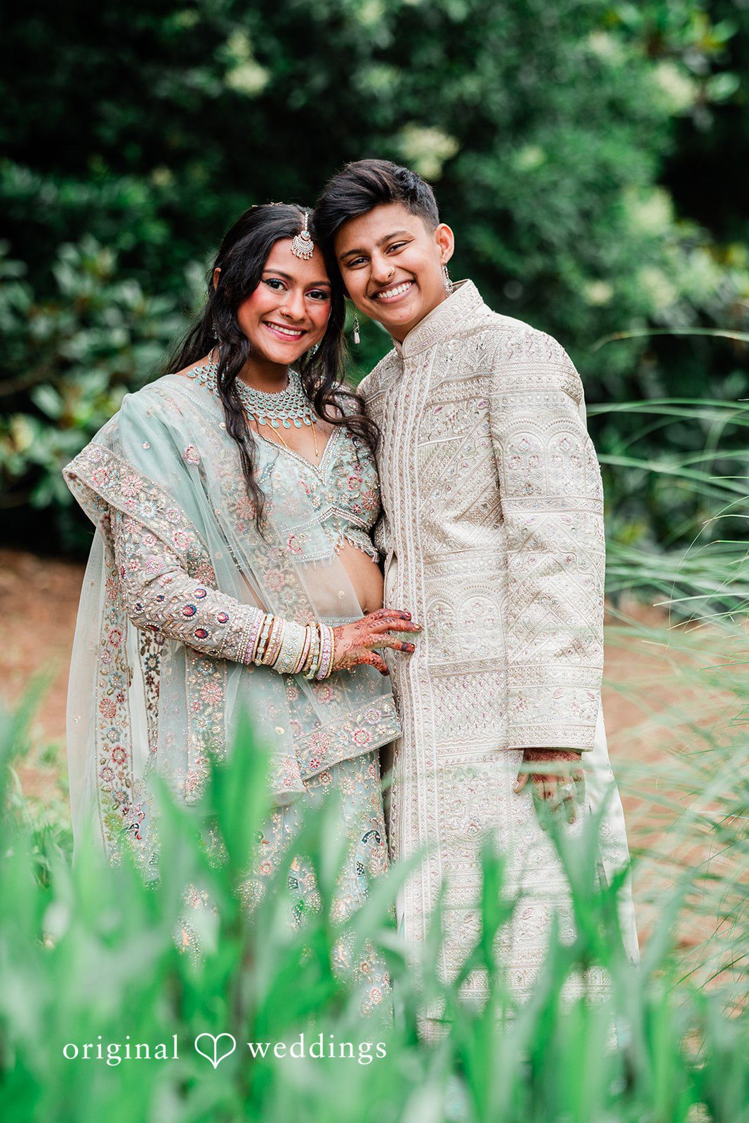 Sonya + Akshaya A happy bride and groom posing closely together amidst green foliage.