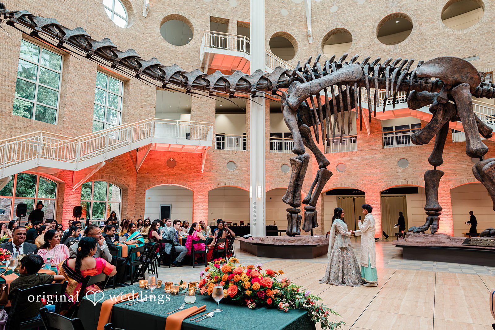 Sonya + Akshaya A wedding ceremony taking place in a large hall beneath a massive dinosaur skeleton exhibit.