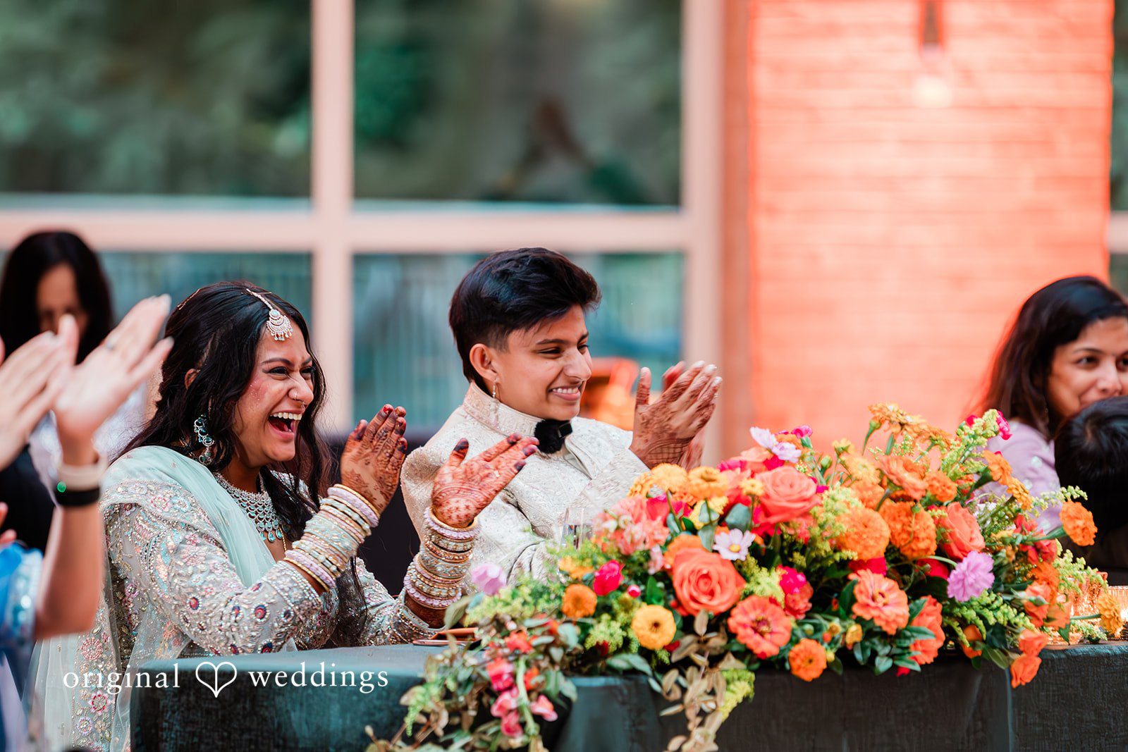 Sonya + Akshaya A couple sitting at a decorated table laughing and clapping with wedding guests.