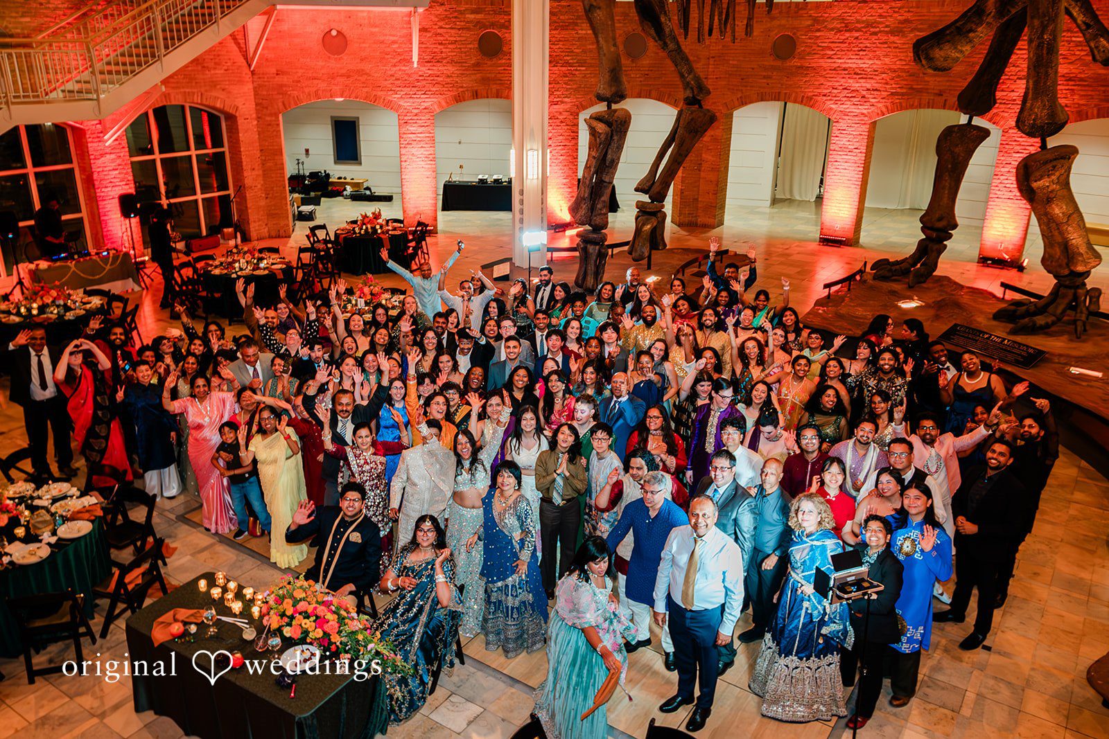 Sonya + Akshaya A large group photo of wedding guests waving from an expansive reception hall floor.