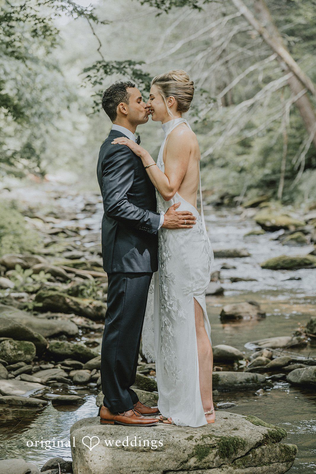 Bride and groom embracing and sharing a kiss by a shallow rocky river.