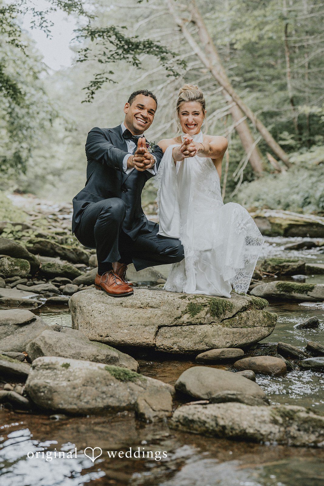 Bride and groom sitting playfully on river stones while holding hands.