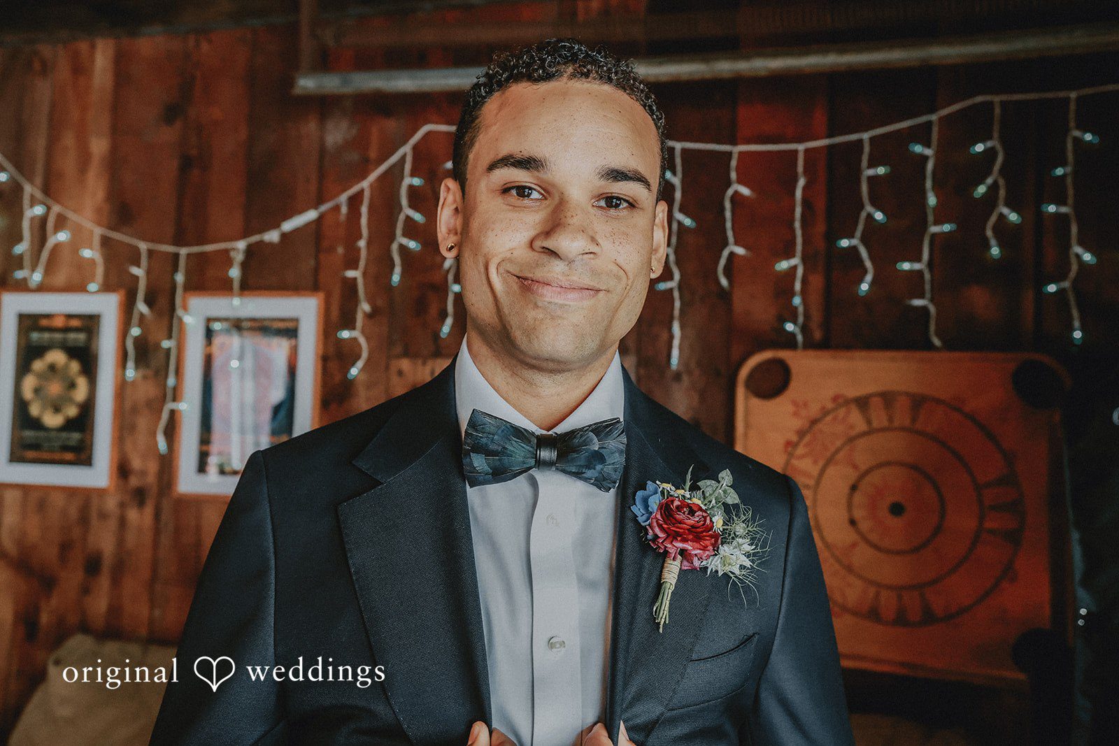 Groom in a dark suit and bow tie smiling in front of a rustic wooden backdrop.