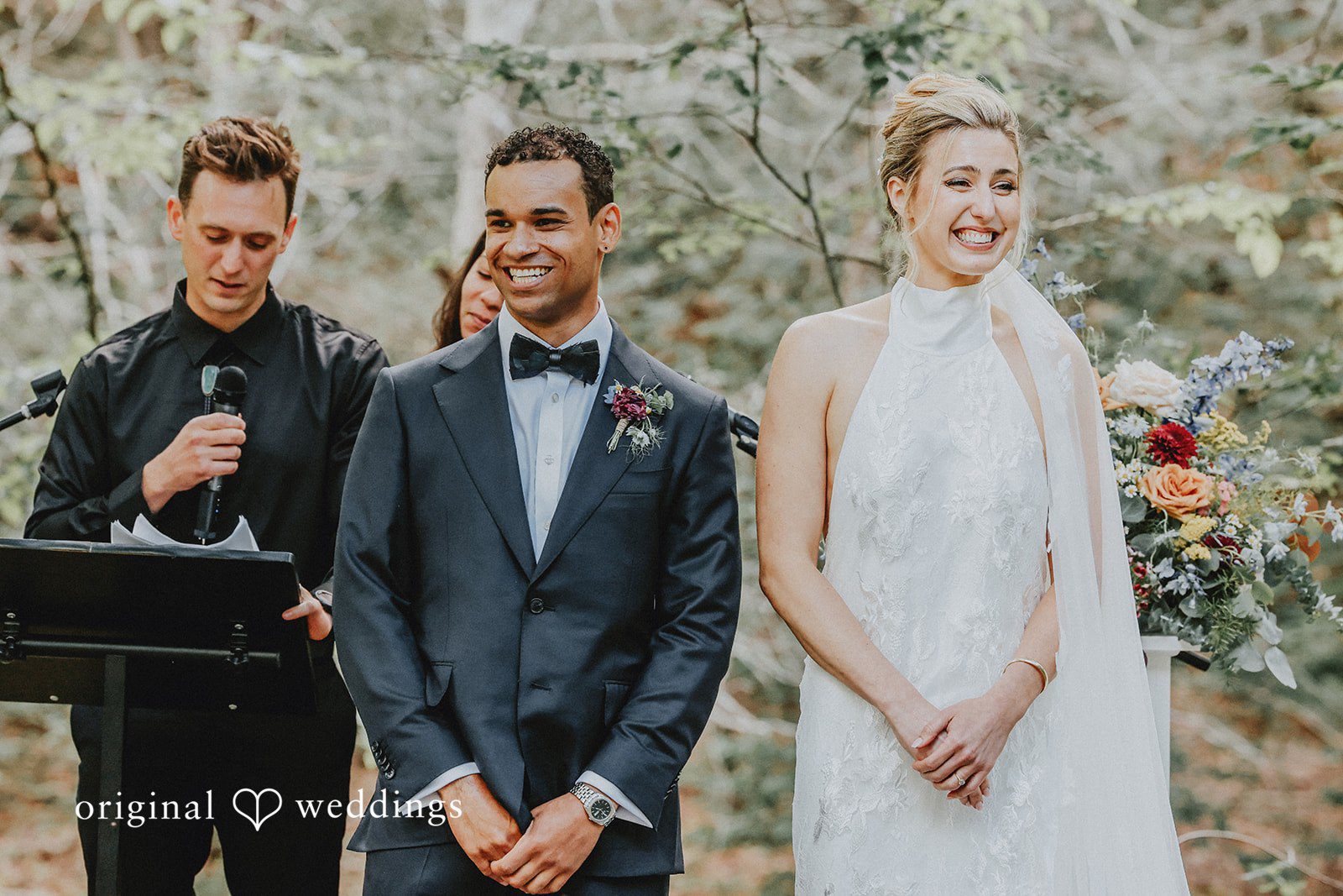 Bride and groom standing together smiling while an officiant speaks into a microphone.