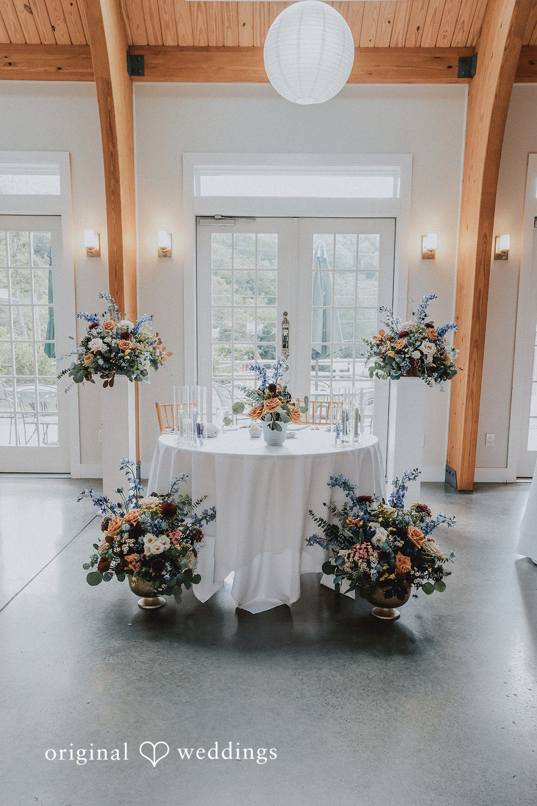 Round reception table dressed in white linen with floral arrangements on a concrete floor.