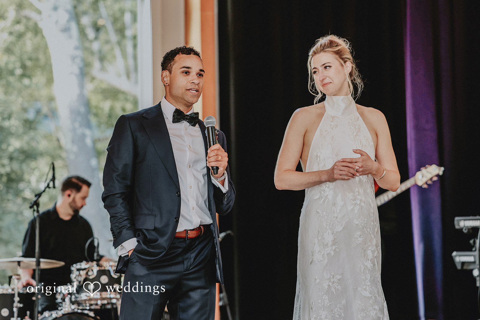 Groom speaking into a microphone while the bride stands smiling next to him.