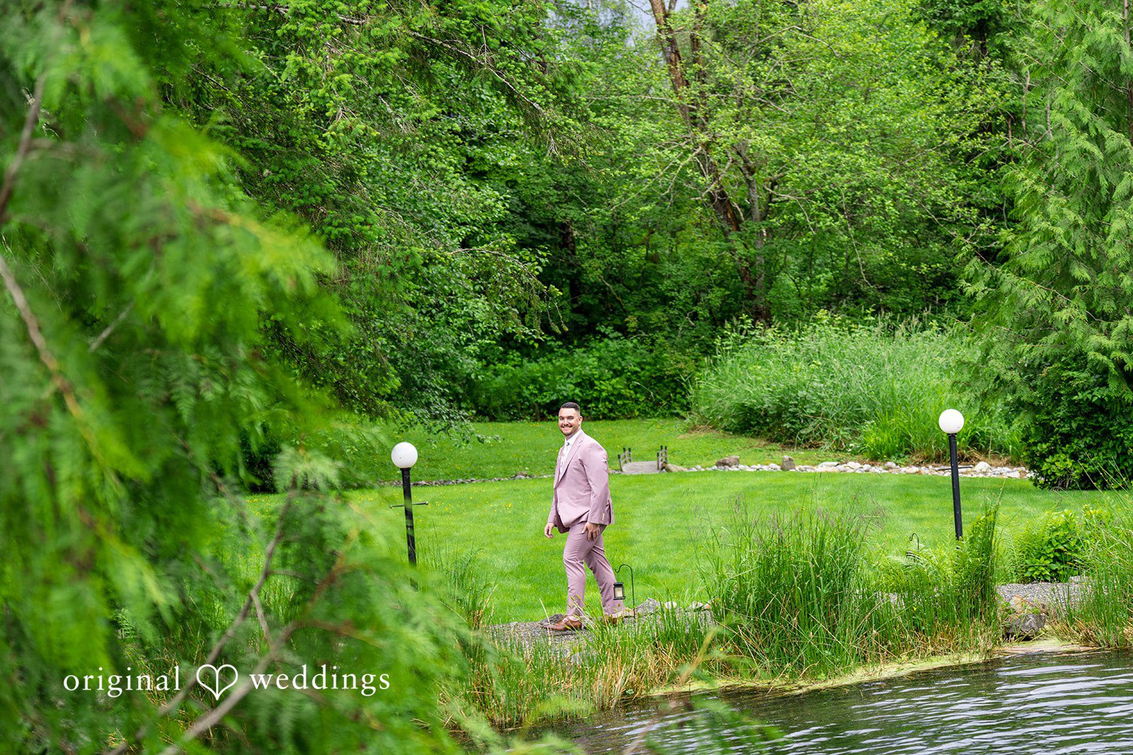 Kristian + Jeff A man in a mauve suit standing along a grassy path near water and dense green trees.