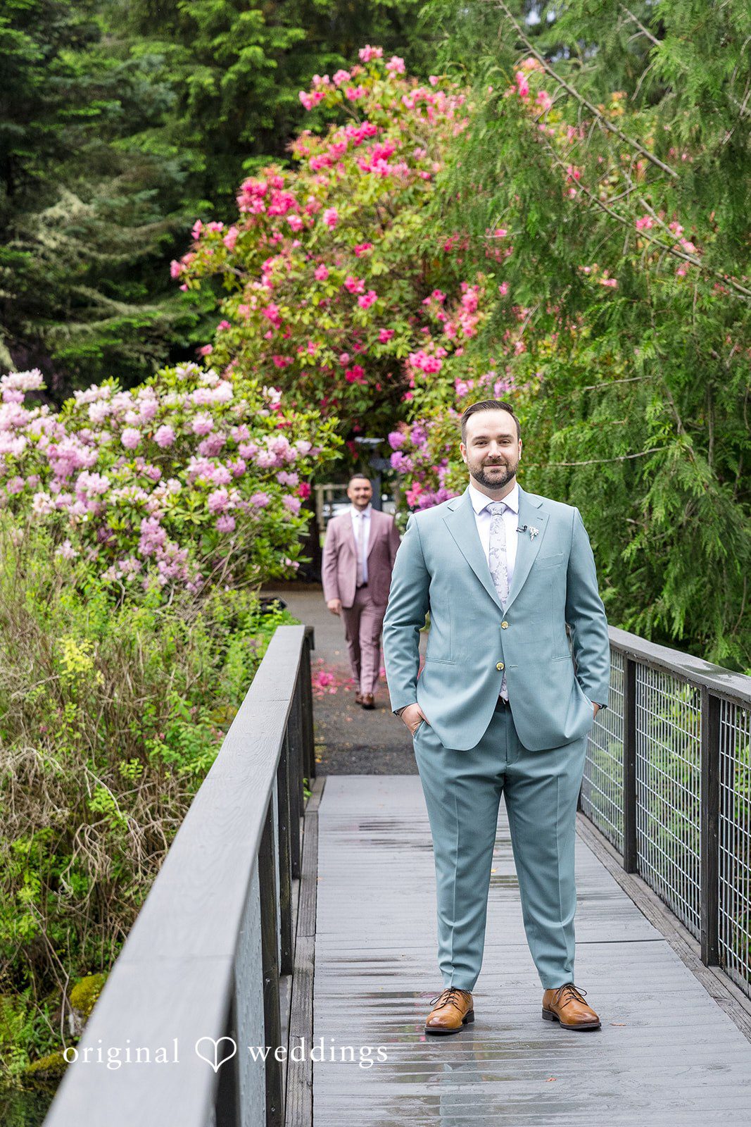 Kristian + Jeff A groom in a light blue suit standing on a bridge walkway with lush greenery behind him.