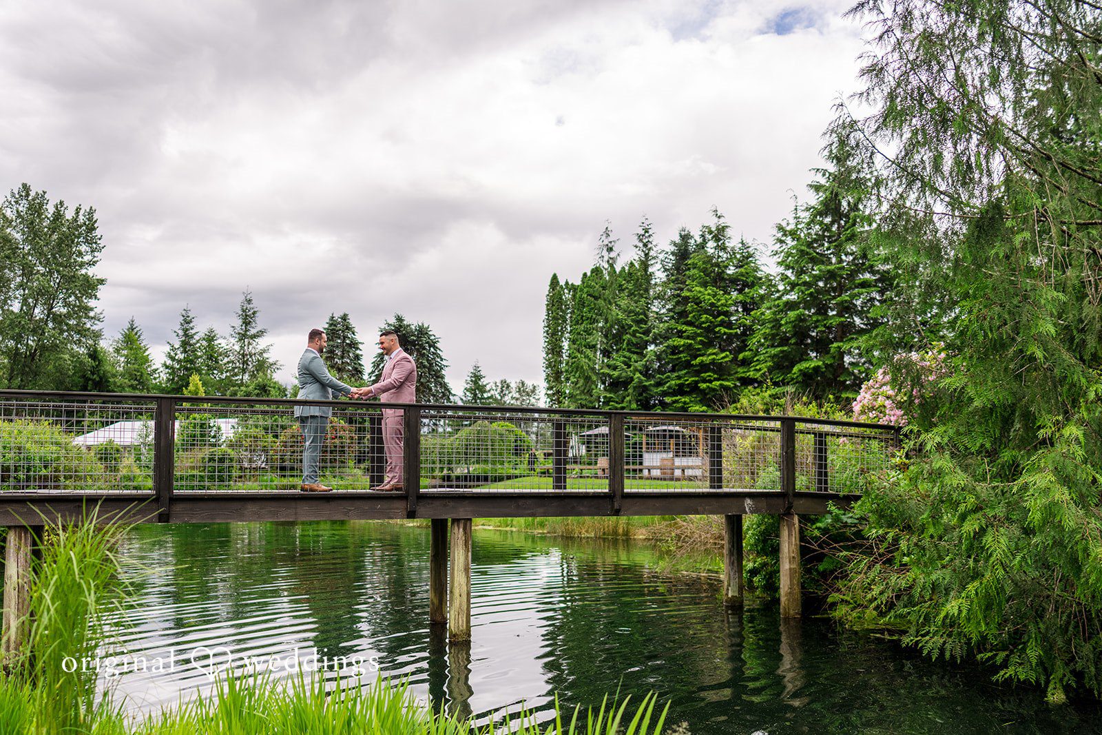 Kristian + Jeff Wide view of couple standing on a wooden footbridge over a scenic pond.