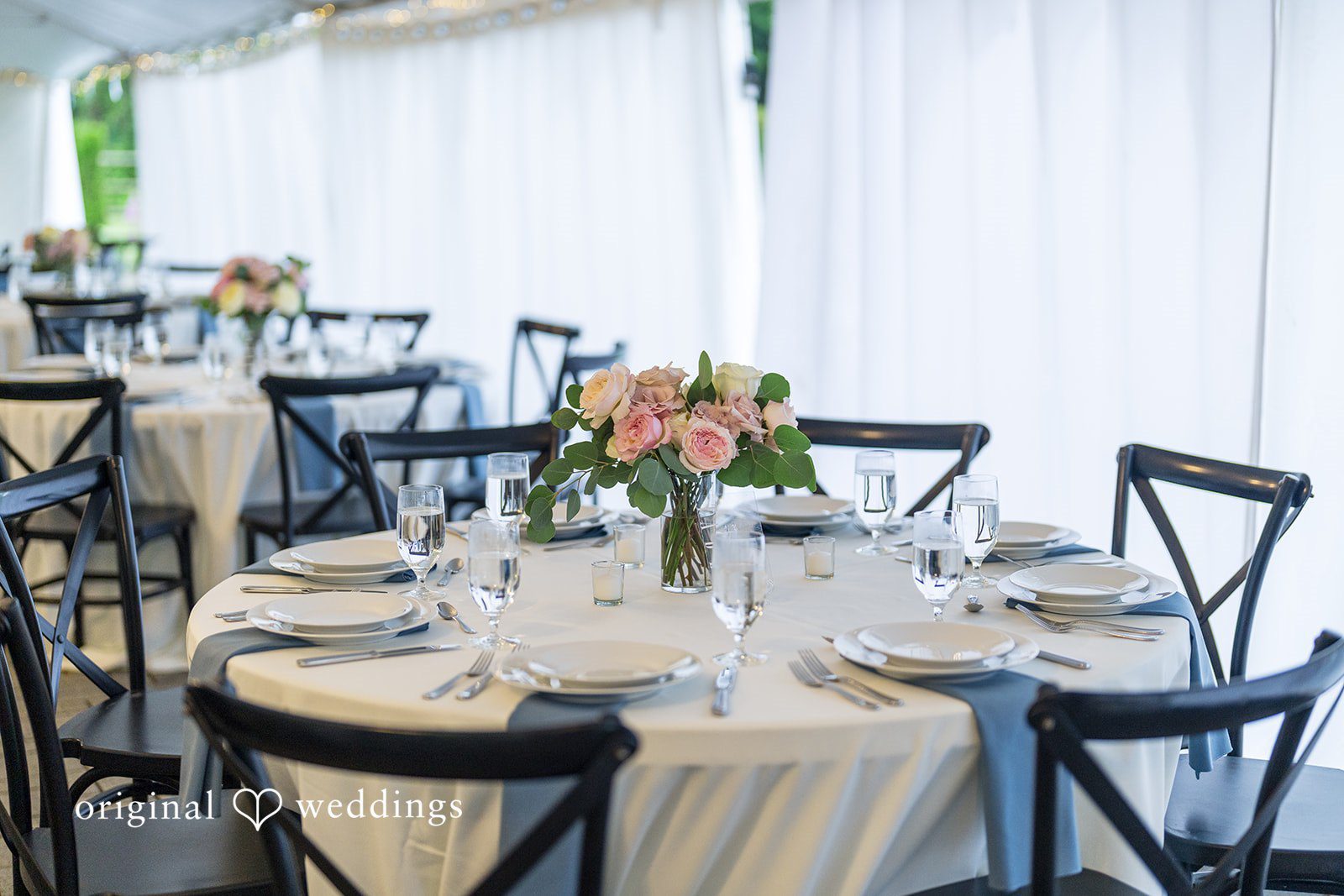Kristian + Jeff Round banquet tables with white tablecloths and dark cross-back chairs arranged for an event.