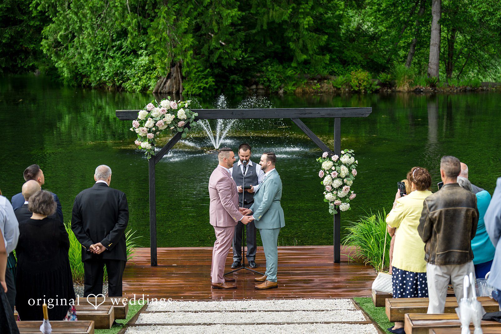 Kristian + Jeff A wedding ceremony under a dark wooden arbor decorated with white flowers, with guests seated on both sides.