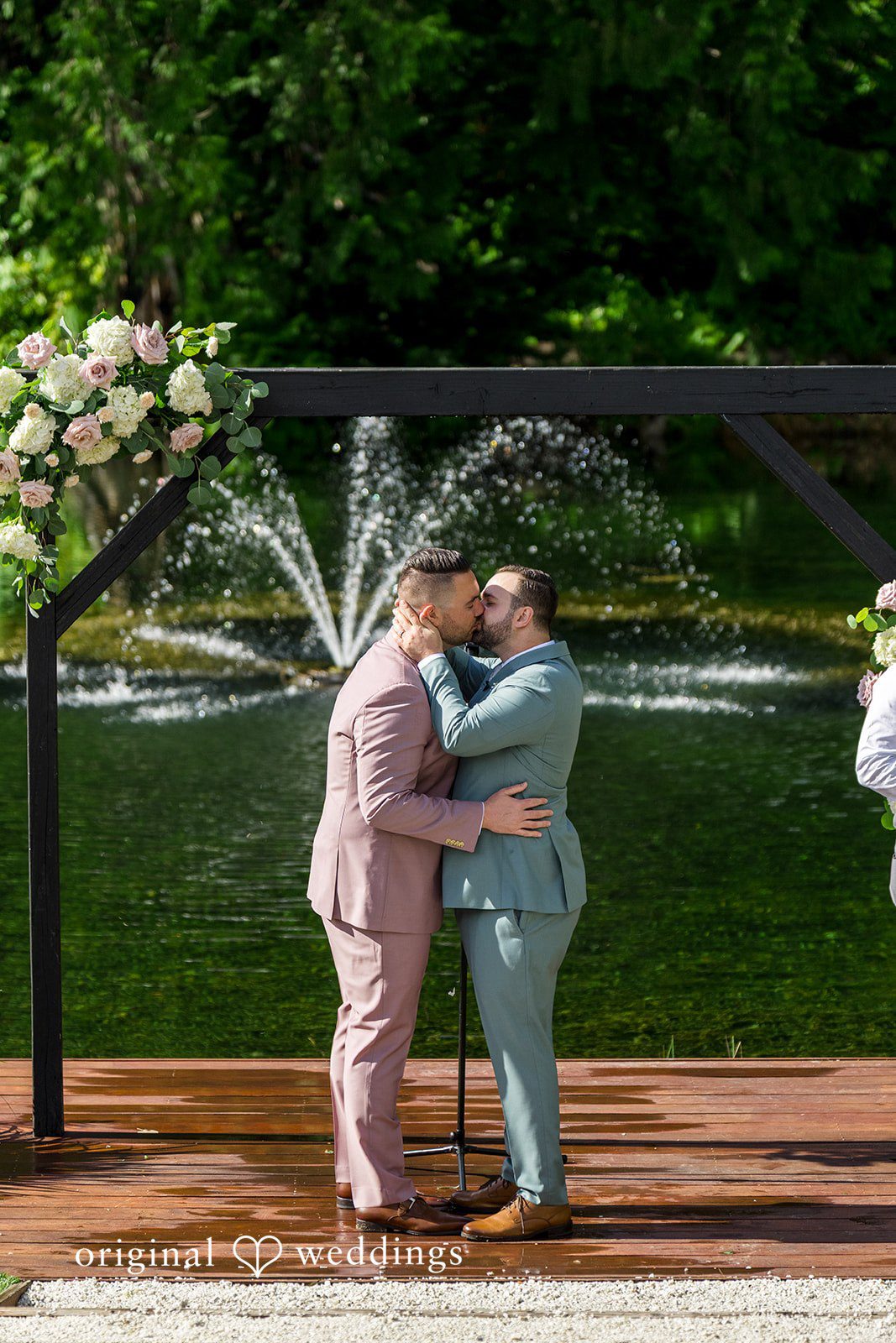 Kristian + Jeff Couple sharing a kiss under a decorated wedding arbor with a water fountain in the background.